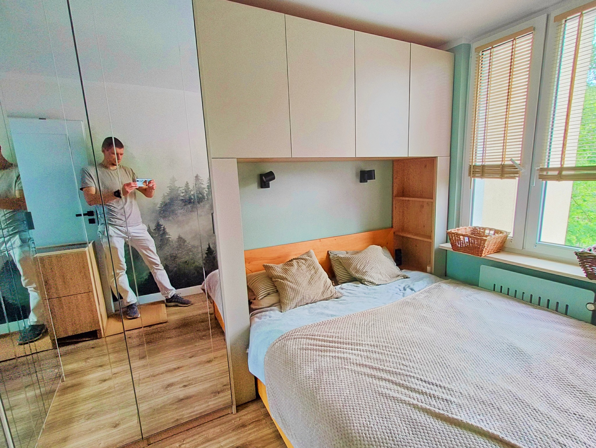 Sypialnia z łóżkiem, szafą wnękową z lustrem i nadstawką szafkową. W lustrze odbija się osoba fotografująca wnętrze. Jasne kolory, naturalne światło.