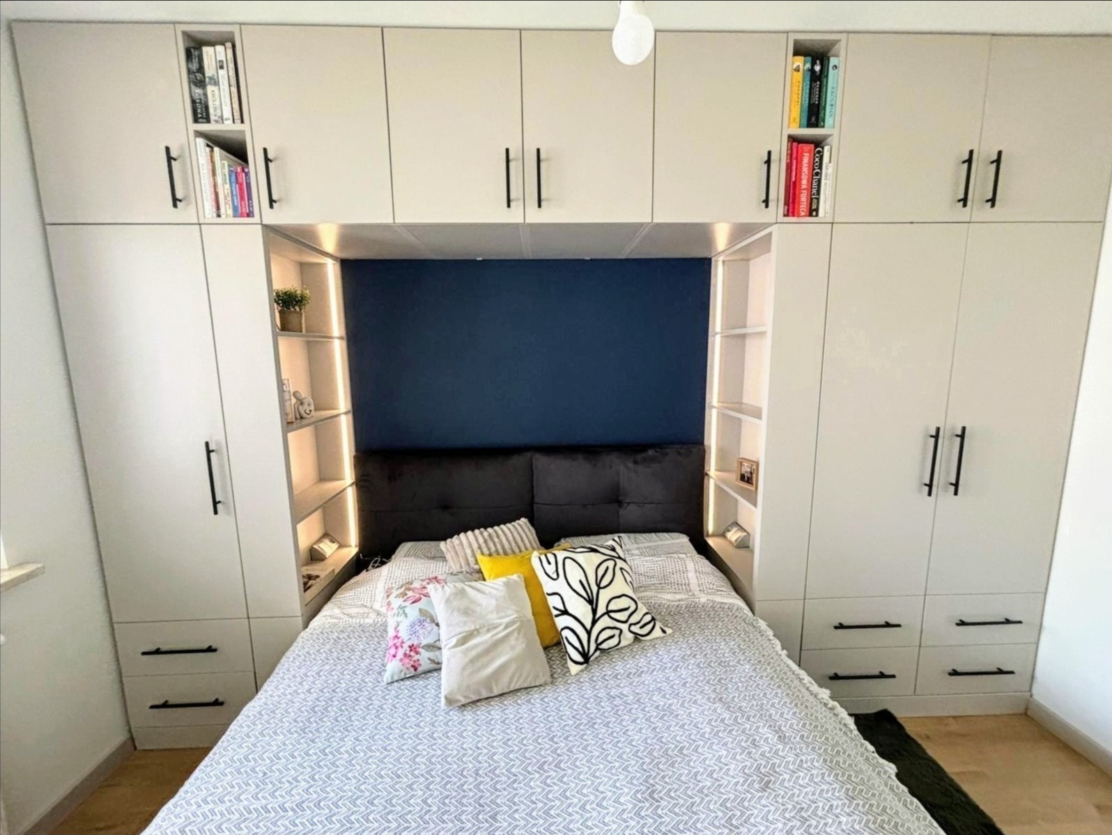 Biała szafa wnękowa z półkami i szafkami nad łóżkiem, czarne uchwyty, oświetlenie LED półek, dekoracyjne poduszki na łóżku, granatowa ściana w tle.