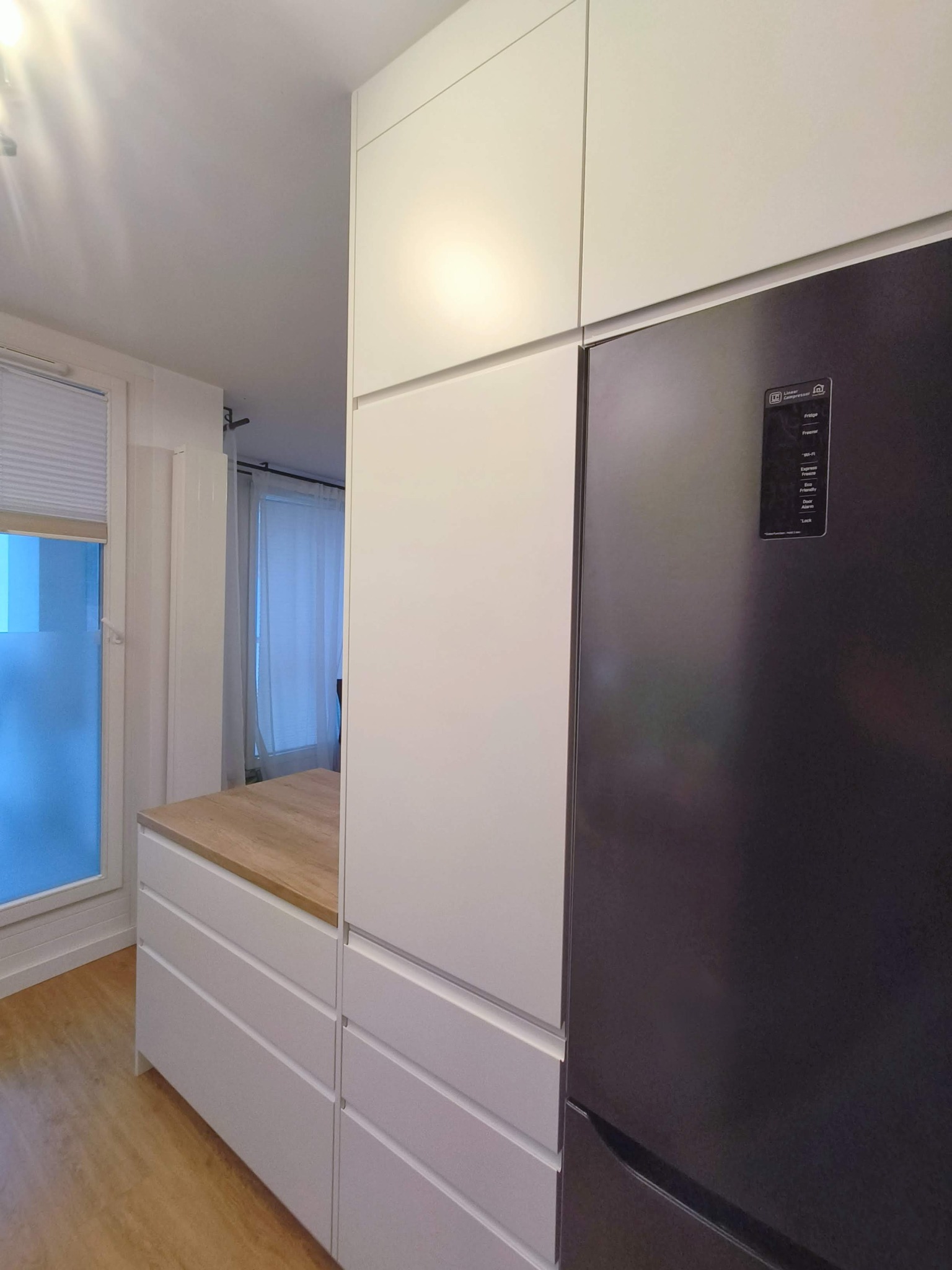 Nowoczesna zabudowa kuchenna z białymi frontami i drewnianym blatem, sąsiadująca z czarną lodówką. Minimalistyczny design i jasne wnętrze.