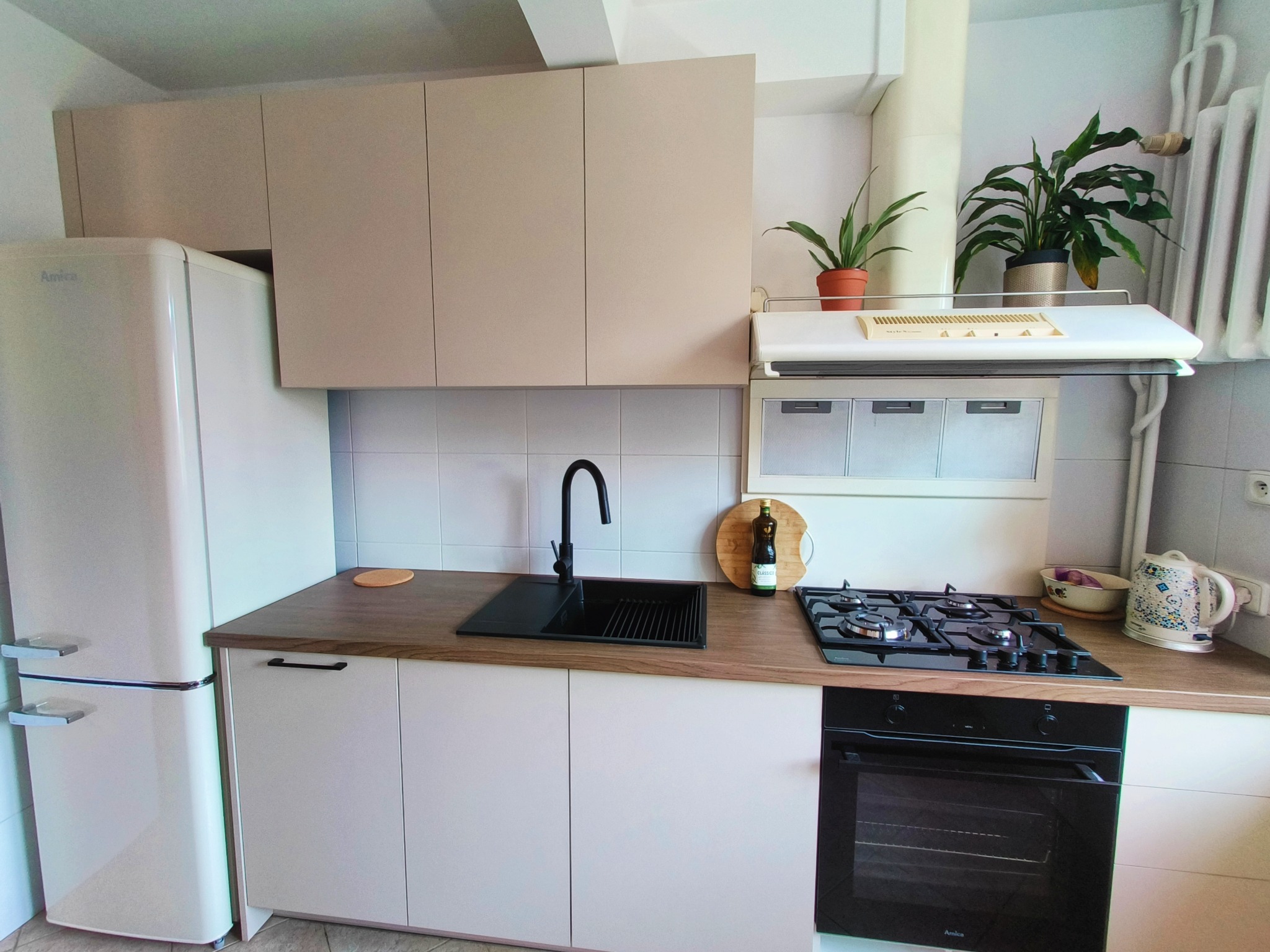 Beżowa zabudowa kuchenna z czarnym zlewem i płytą gazową, uzupełniona o lodówkę Amica w stylu retro i rośliny doniczkowe, tworzy przytulną przestrzeń.