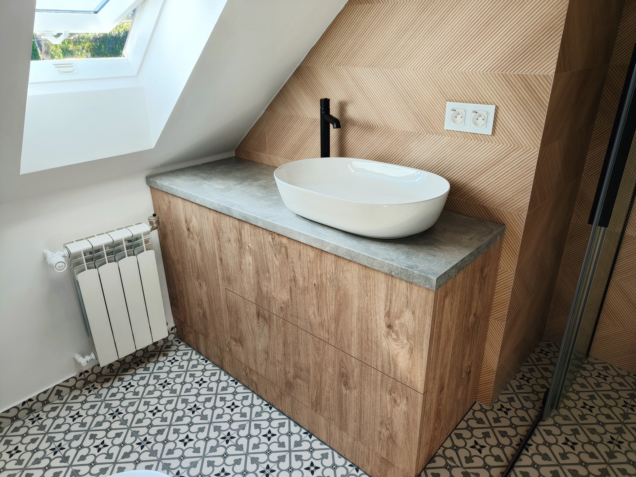 Nowoczesna łazienka na poddaszu z drewnianą szafką pod umywalkę, owalną białą umywalką nablatową i czarną armaturą. Uwagę zwraca wzorzysta podłoga i skośny sufit.