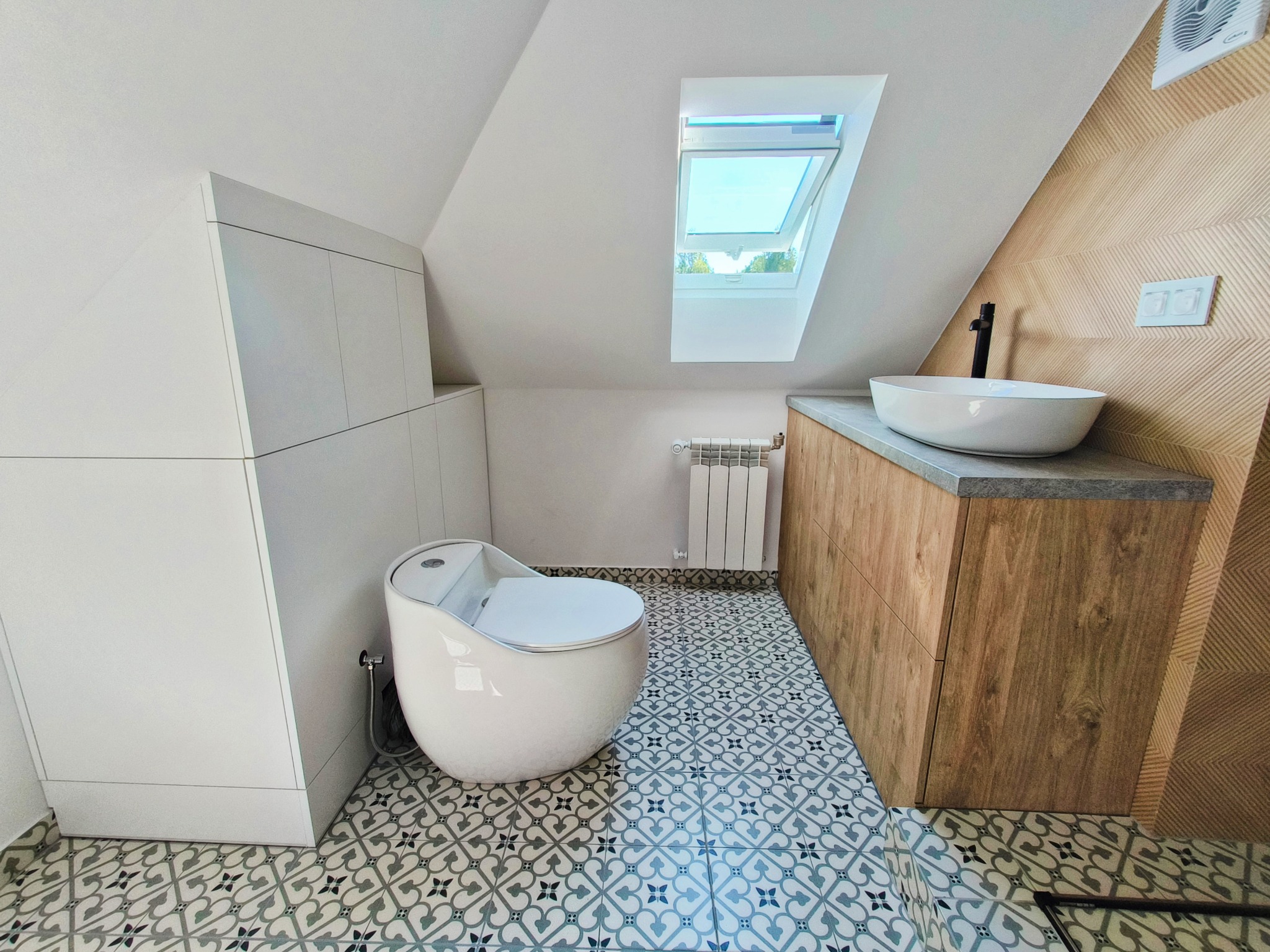 Aranżacja łazienki na poddaszu ze skośnym oknem dachowym, białą ceramiką i szafką z drewnianym blatem. Geometryczne płytki podłogowe.