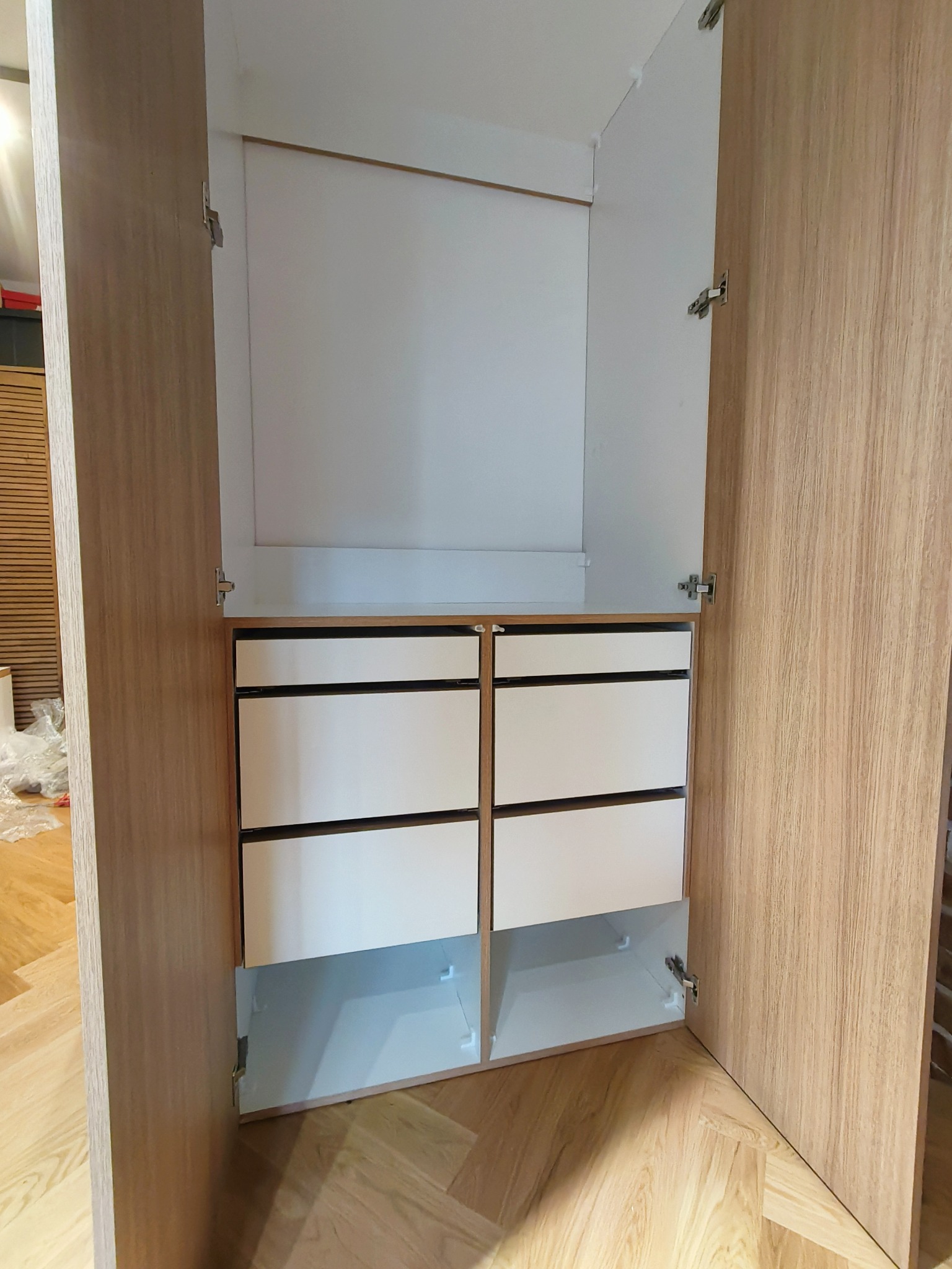 Otwarta szafa z drewnopodobnymi drzwiami i białymi szufladami. Widoczne okucia i wnętrze szafy, podłoga z paneli. Ujęcie z bliska, wnętrze szafy dobrze oświetlone.