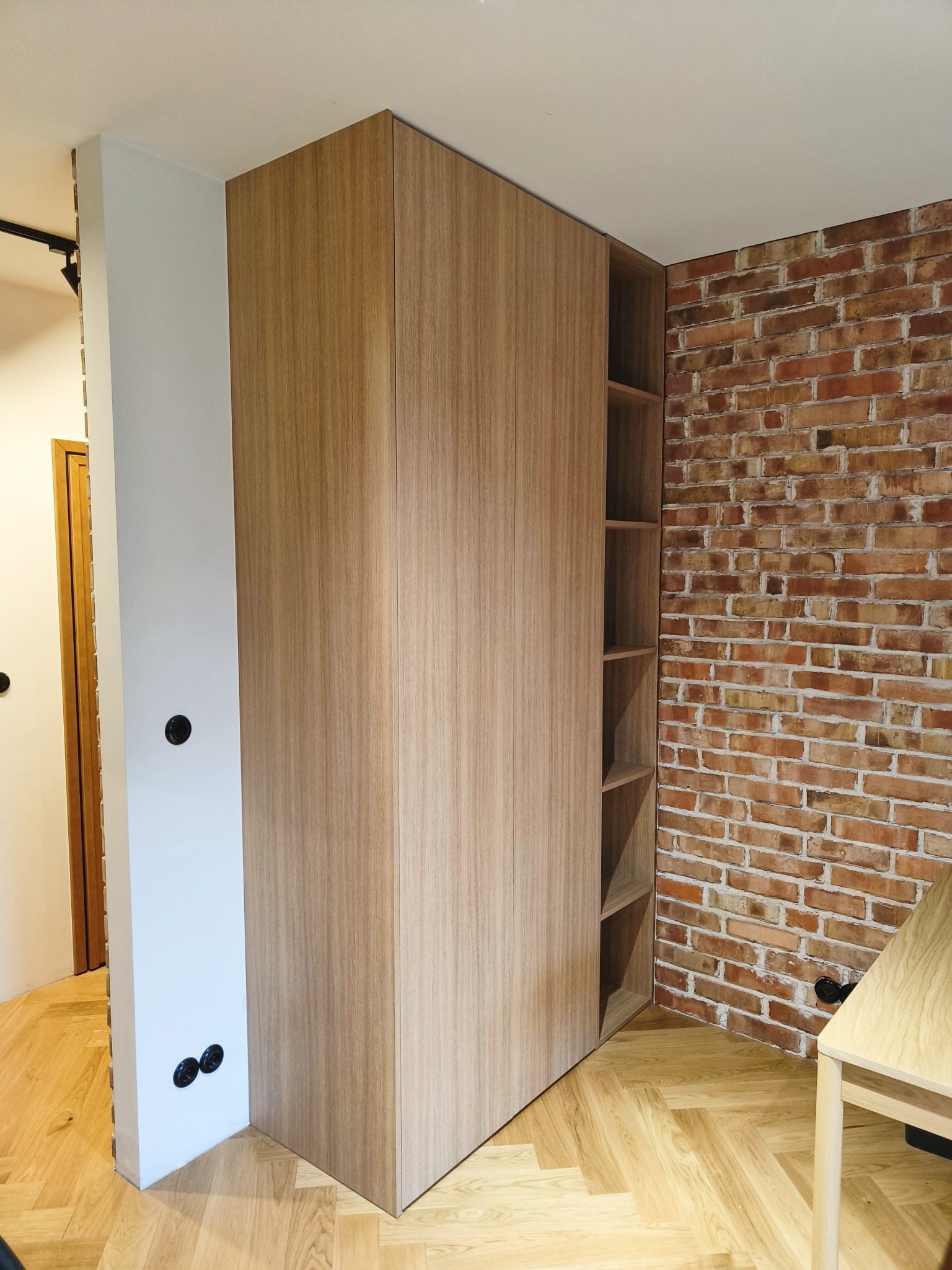 Drewniana szafa wnękowa z otwartymi półkami, zlicowana ze ścianą, na tle ceglanej ściany i parkietu ułożonego w jodełkę. Minimalistyczny design.