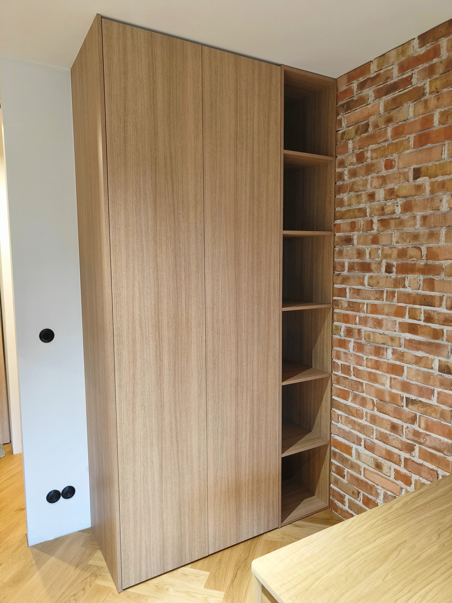 Jasna szafa wnękowa z otwartymi półkami, dopasowana do ceglanej ściany. Minimalistyczny design, widoczne gniazdka elektryczne. Podłoga parkiet.