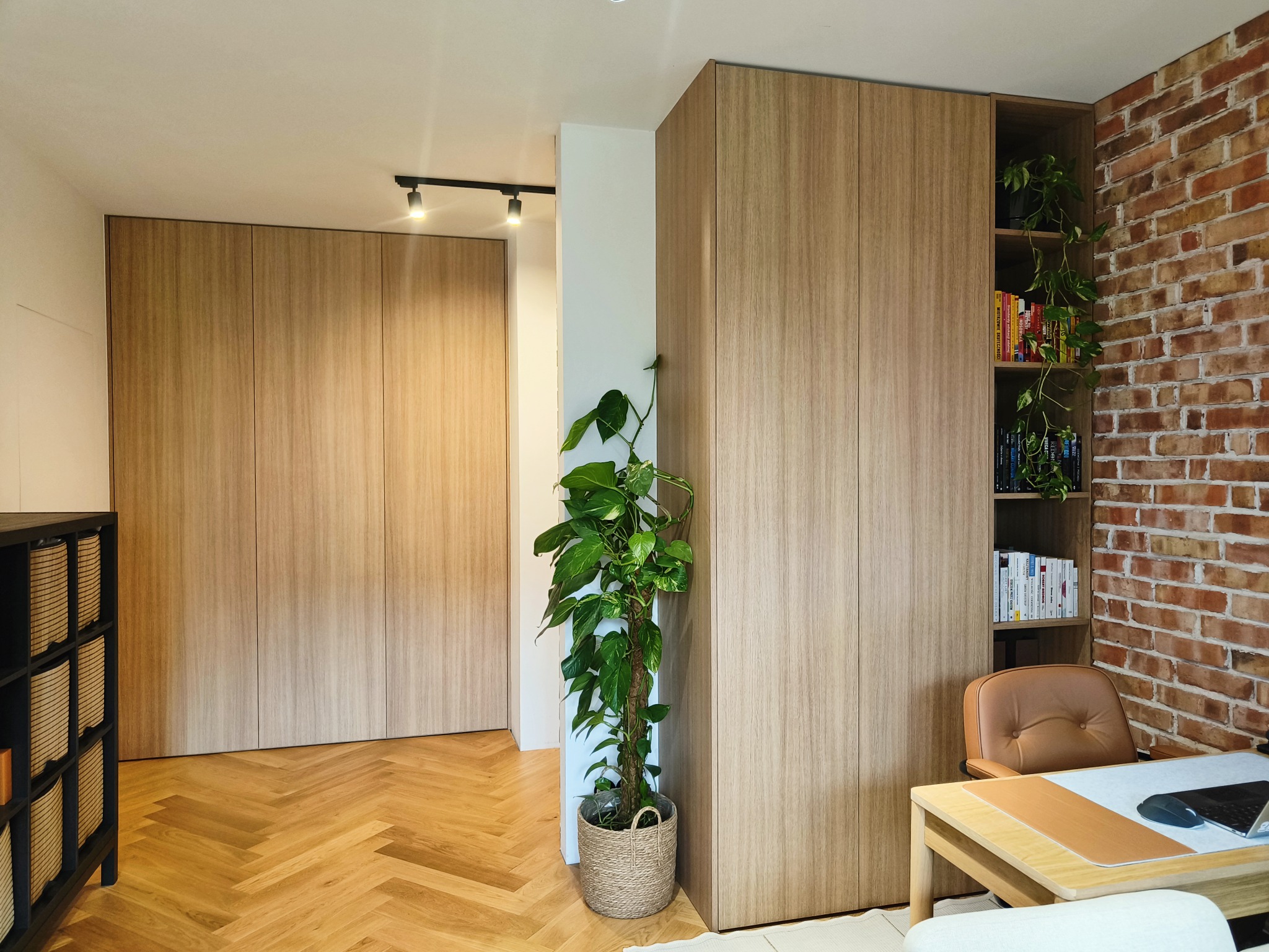 Jasne, drewniane szafy na wymiar w nowoczesnym wnętrzu z parkietem w jodełkę, rośliną doniczkową i ścianą z cegły. Minimalistyczny design z subtelnym oświetleniem.