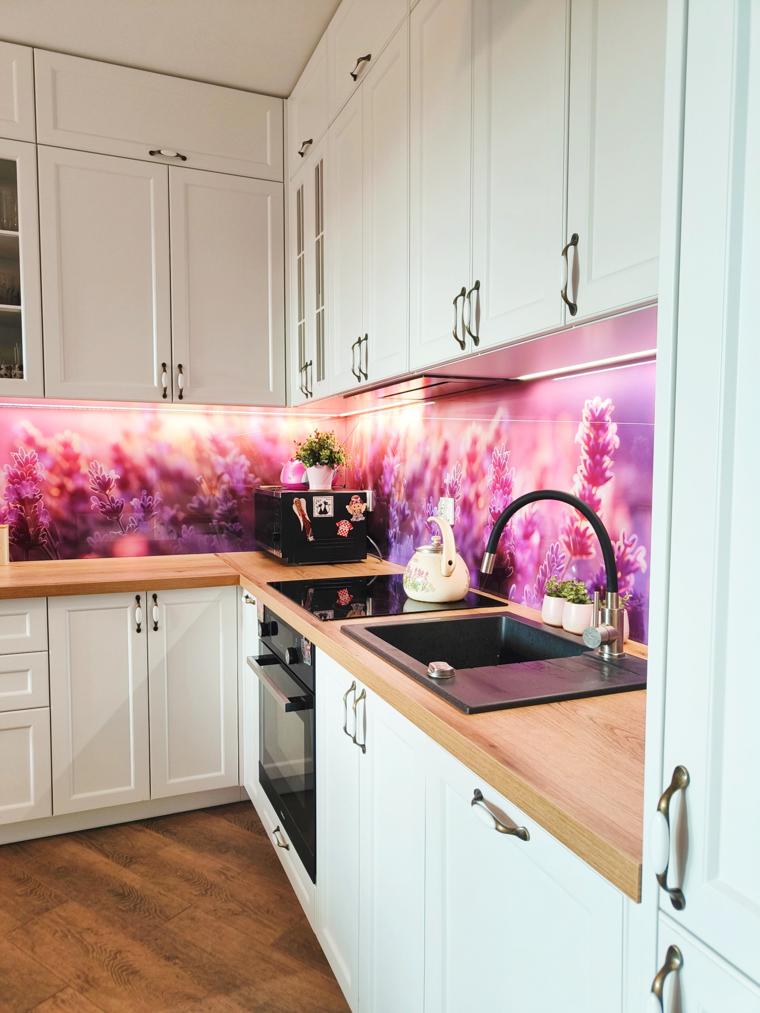 Biała kuchnia z drewnianym blatem i fioletowym panelem z motywem lawendy. Zlewozmywak z czarną baterią, piekarnik w zabudowie. Uwagę zwracają ozdobne uchwyty.