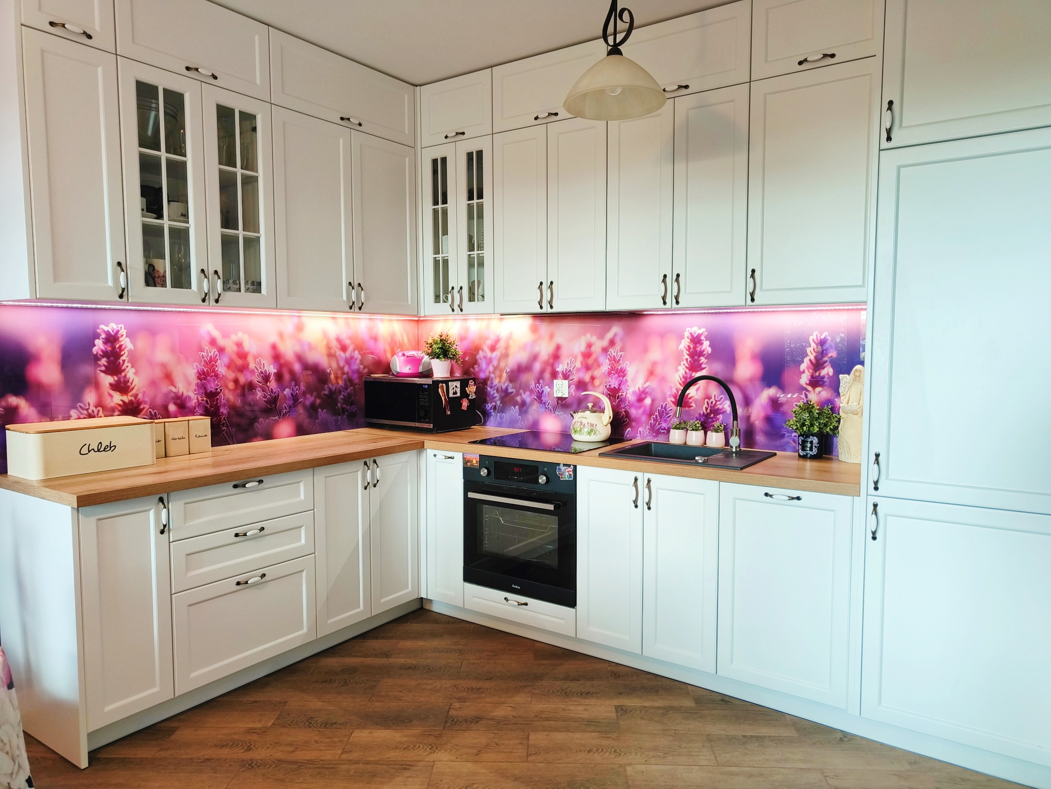 Biała kuchnia z drewnianym blatem, ozdobnym panelem ściennym z motywem lawendy i klasycznymi uchwytami. Widoczny piekarnik i akcesoria kuchenne.