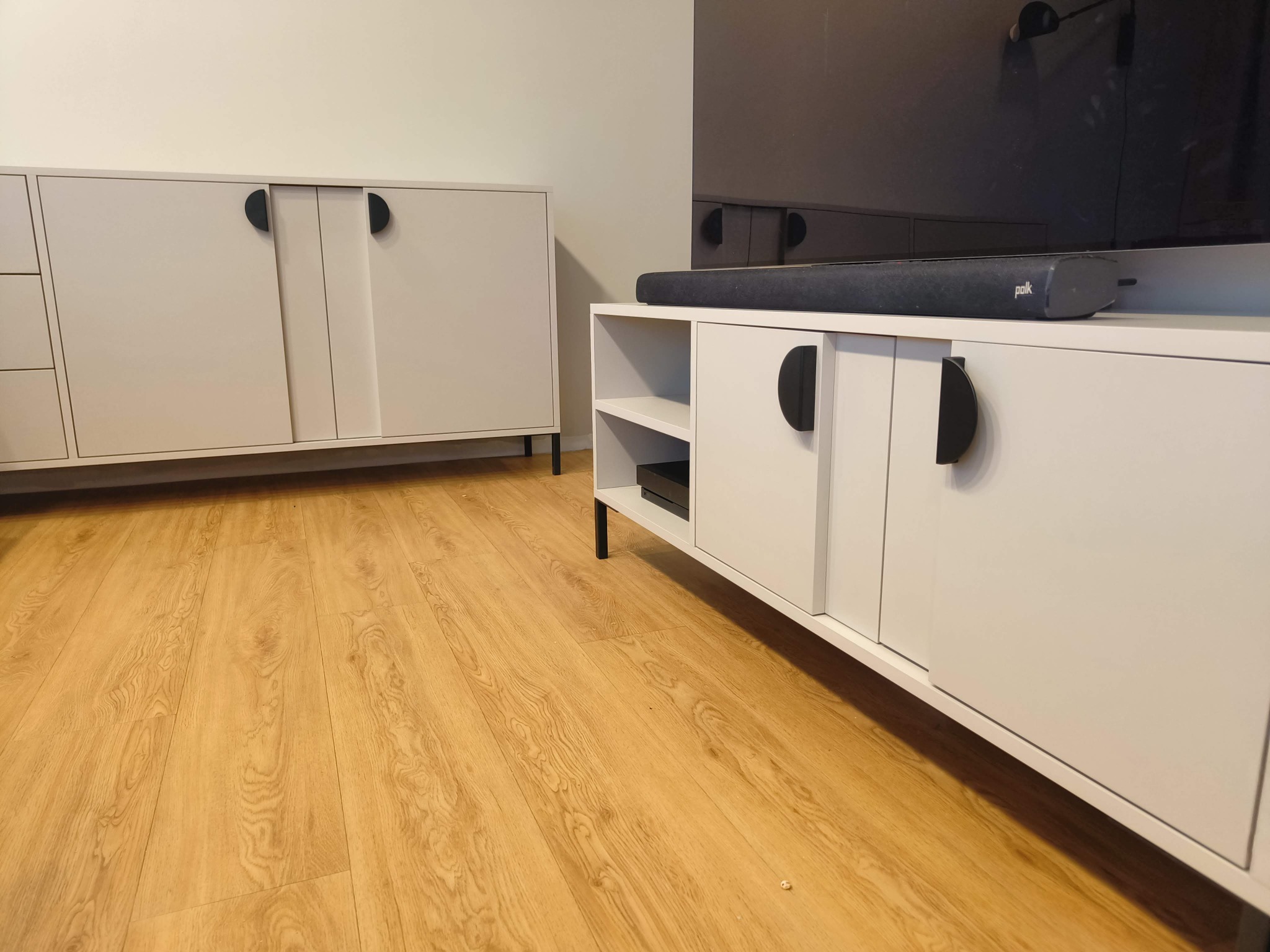 Jasne, minimalistyczne meble RTV z półokrągłymi uchwytami, ustawione na drewnianej podłodze. Widoczny soundbar na szafce i fragment drugiego mebla w tle.