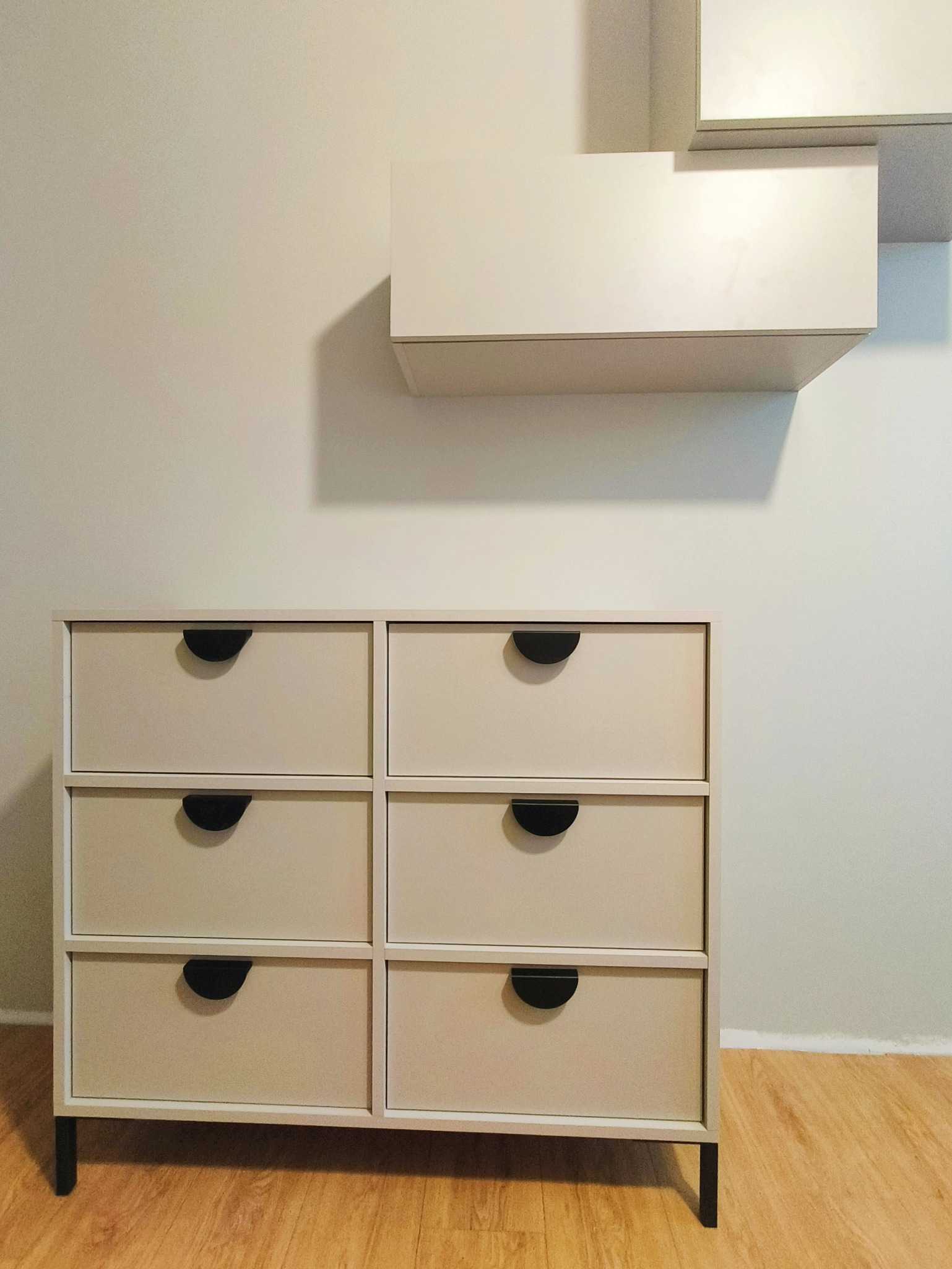 Beżowa komoda z sześcioma szufladami i czarnymi uchwytami, uzupełniona wiszącą szafką na ścianie. Minimalistyczny design, jasne wnętrze.