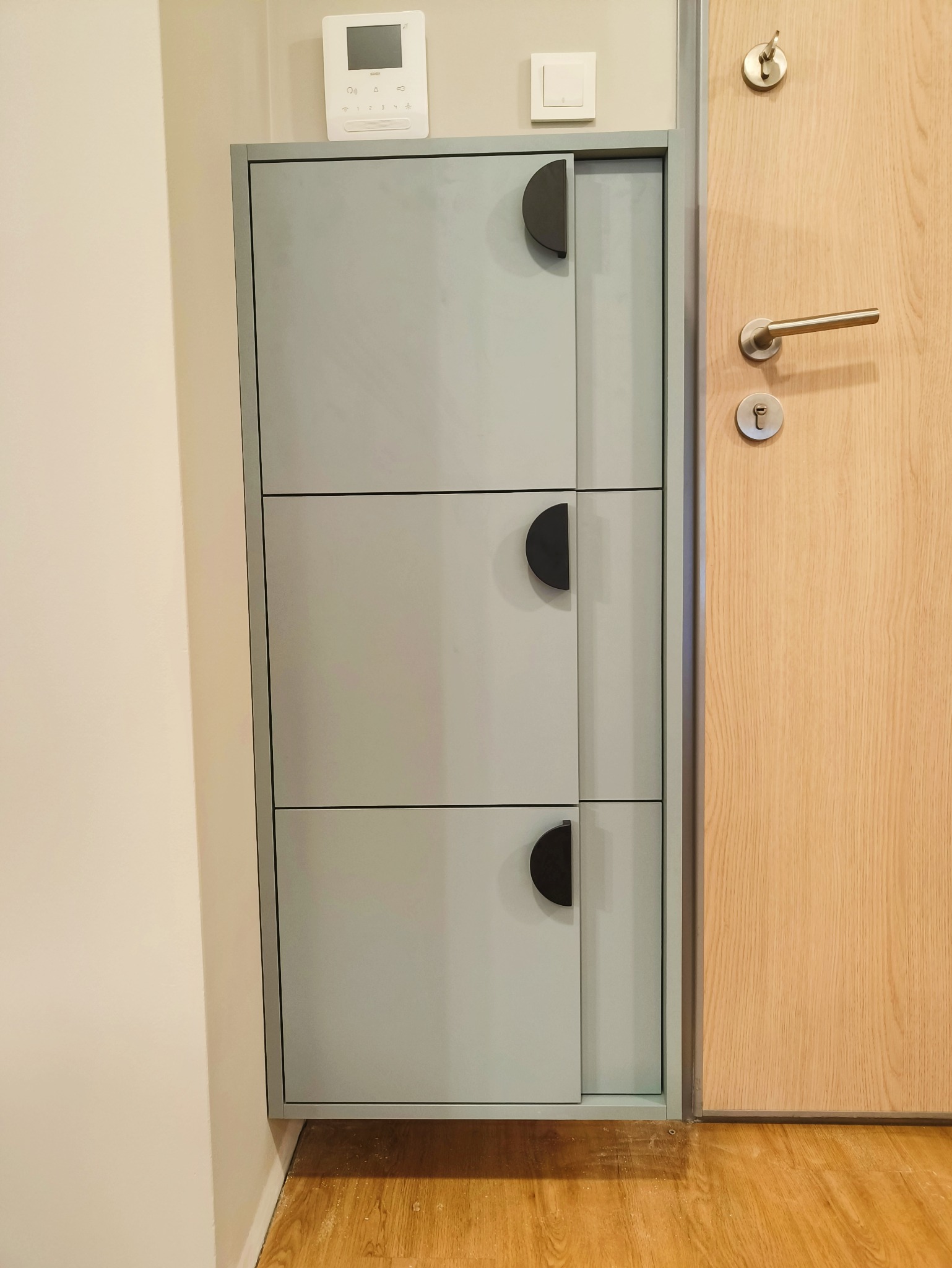 Szara szafka na buty z trzema poziomami i półokrągłymi uchwytami, zamontowana obok drewnianych drzwi wejściowych. Minimalistyczny design, jasne wnętrze.