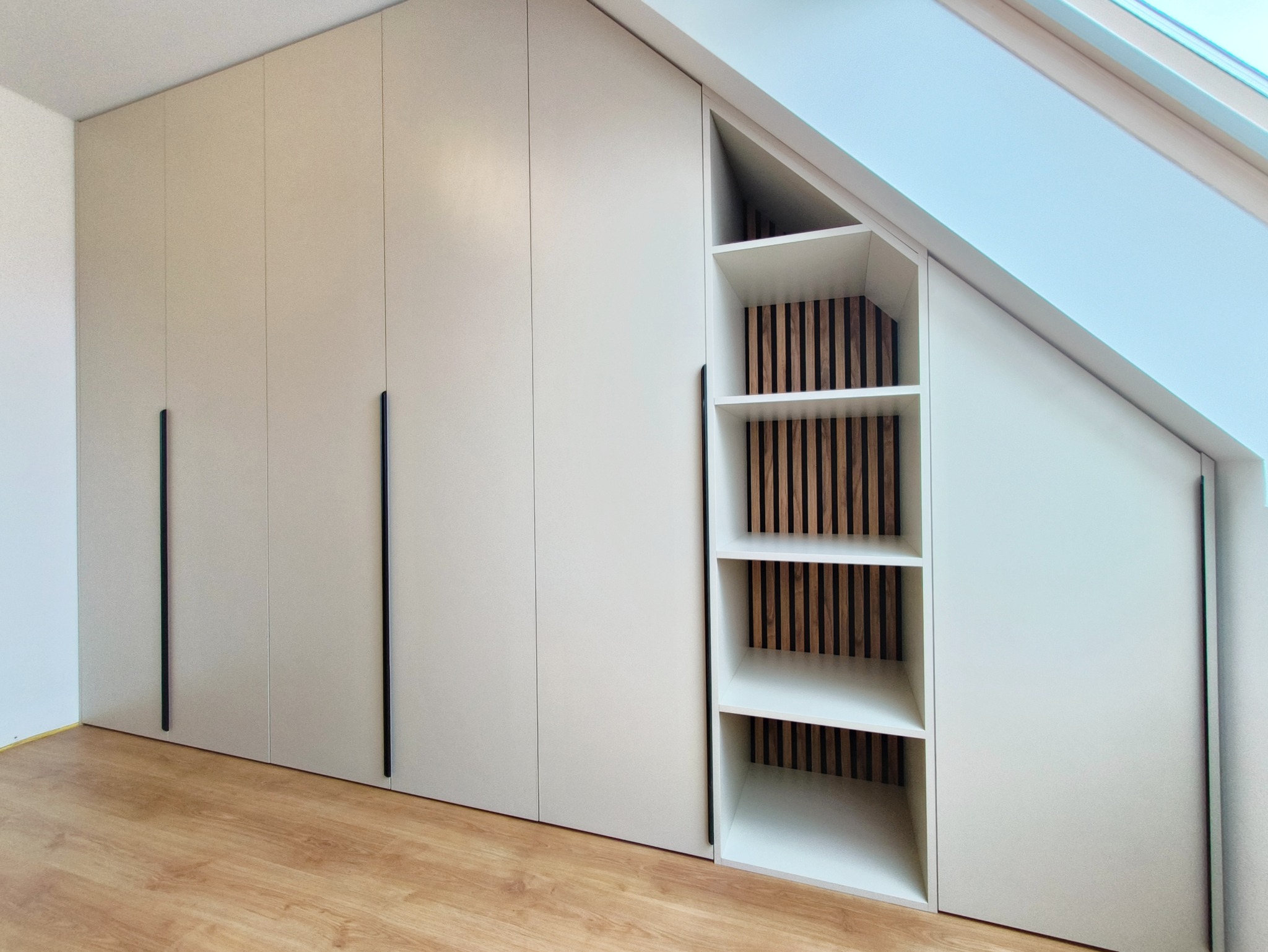 Biała szafa wnękowa z czarnymi uchwytami, dopasowana do skosu dachu, z otwartą półką z drewnianymi lamelami w tle. Nowoczesny design.