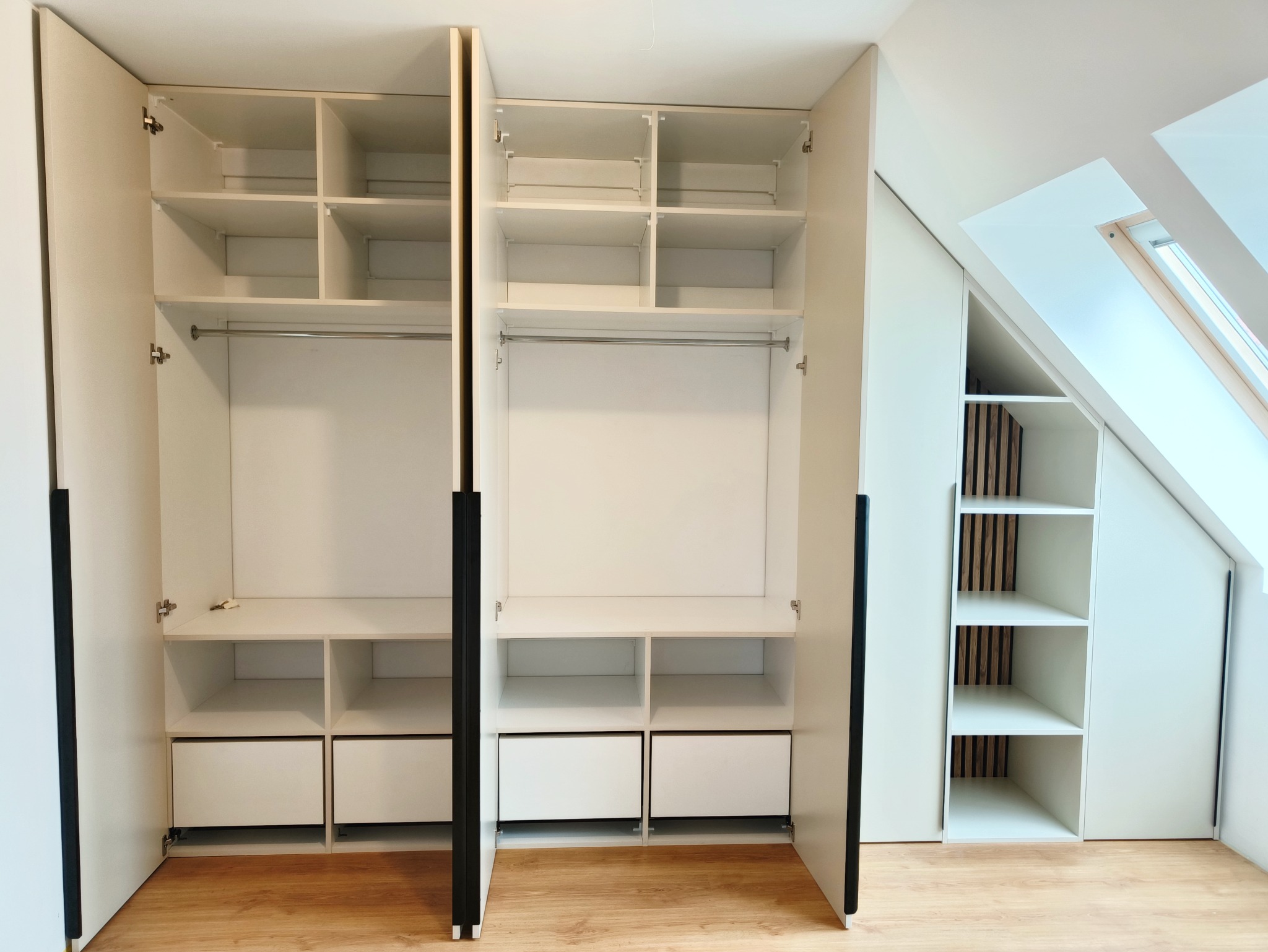 Biała szafa wnękowa z otwartymi drzwiami, widoczne półki, szuflady i drążek na ubrania. Dodatkowa szafka w skosie pod oknem dachowym. Jasne drewno na podłodze.