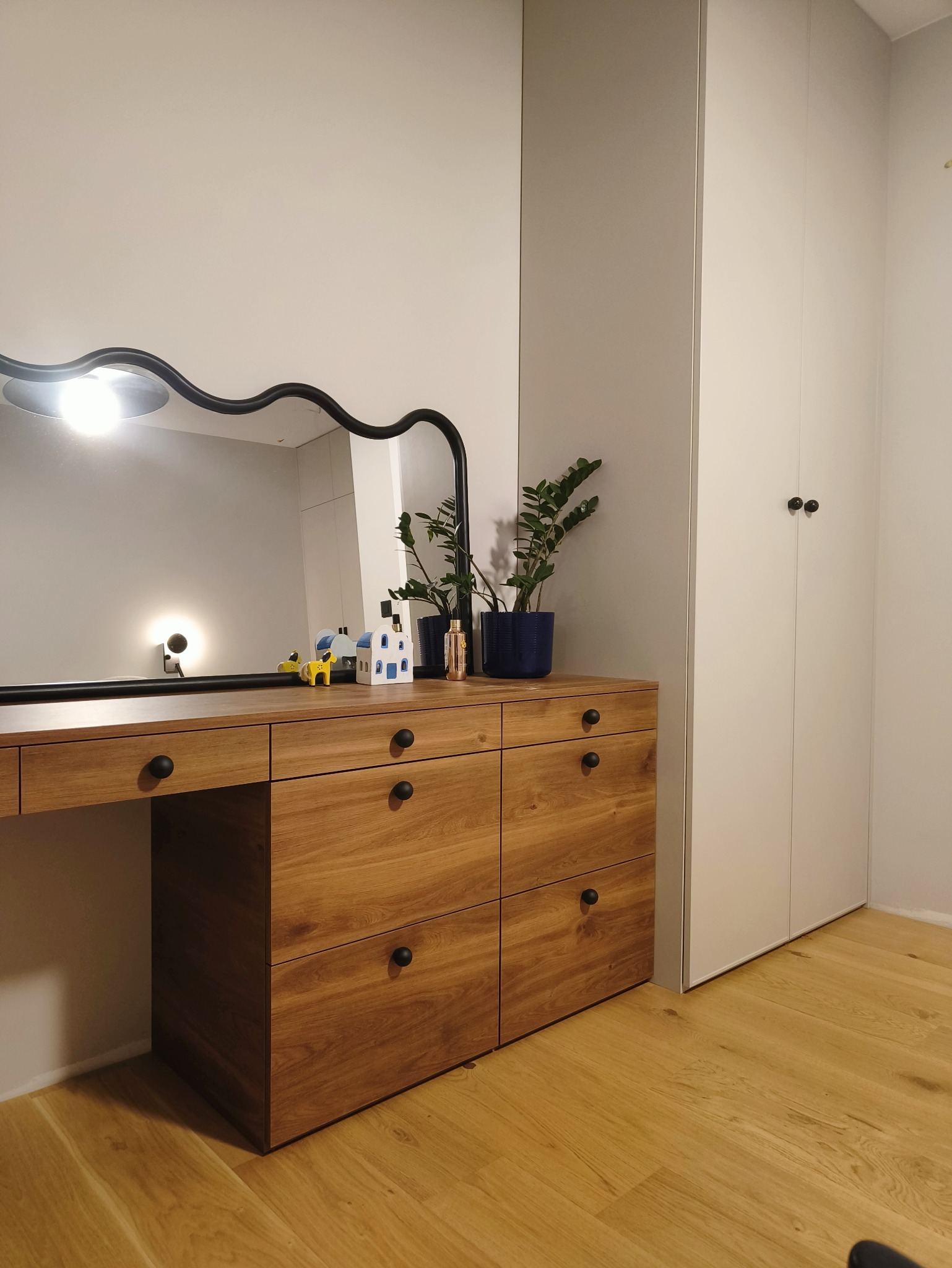Drewniana toaletka z lustrem o falistym obramowaniu i ciemnymi uchwytami szuflad. Obok biała szafa wnękowa z minimalistycznymi gałkami. Na blacie dekoracje i roślina.