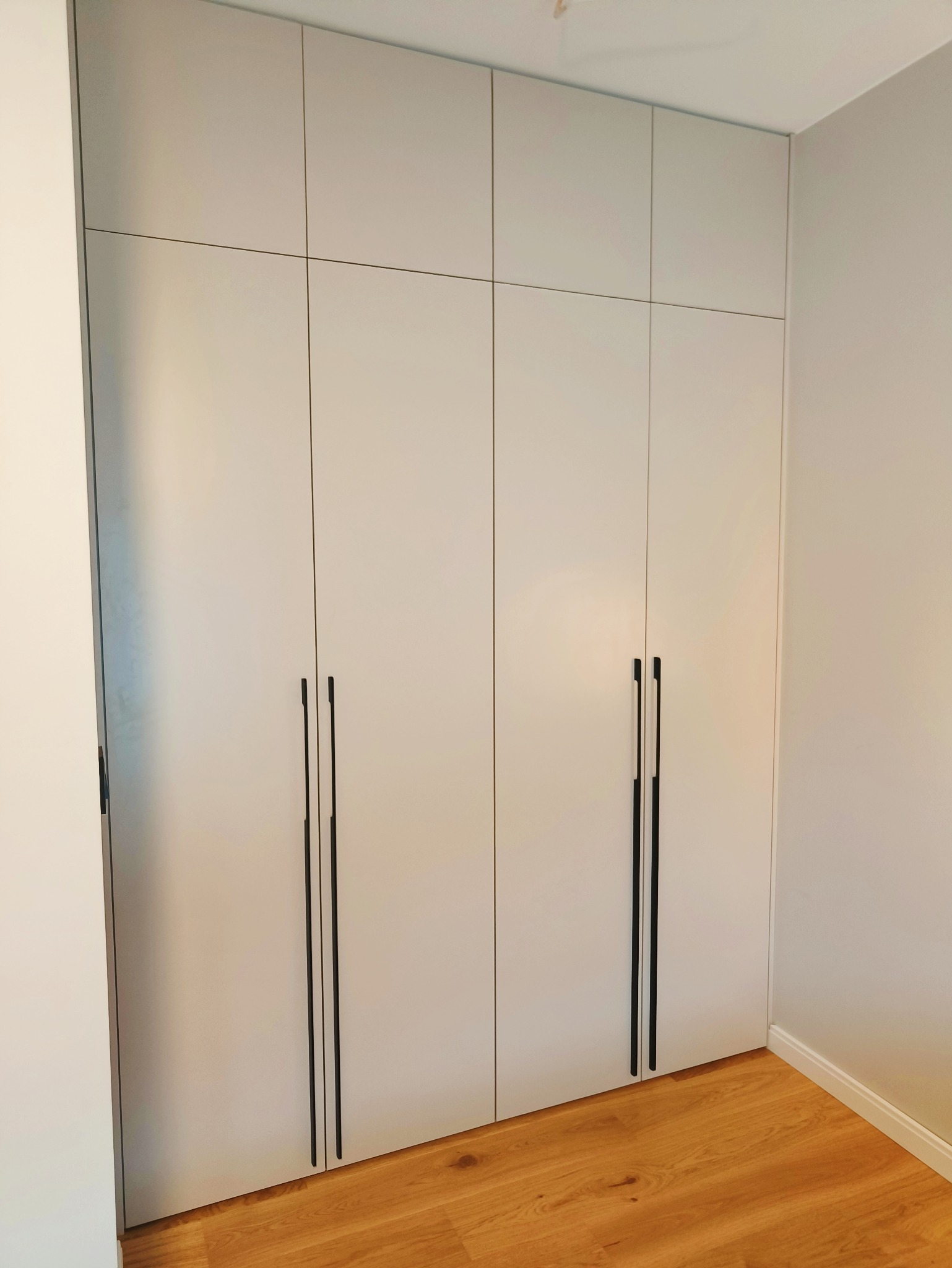 Jasnoszara szafa wnękowa z długimi, czarnymi uchwytami, dopasowana do wnętrza z drewnianą podłogą. Minimalistyczny design, proste linie, nowoczesny styl.