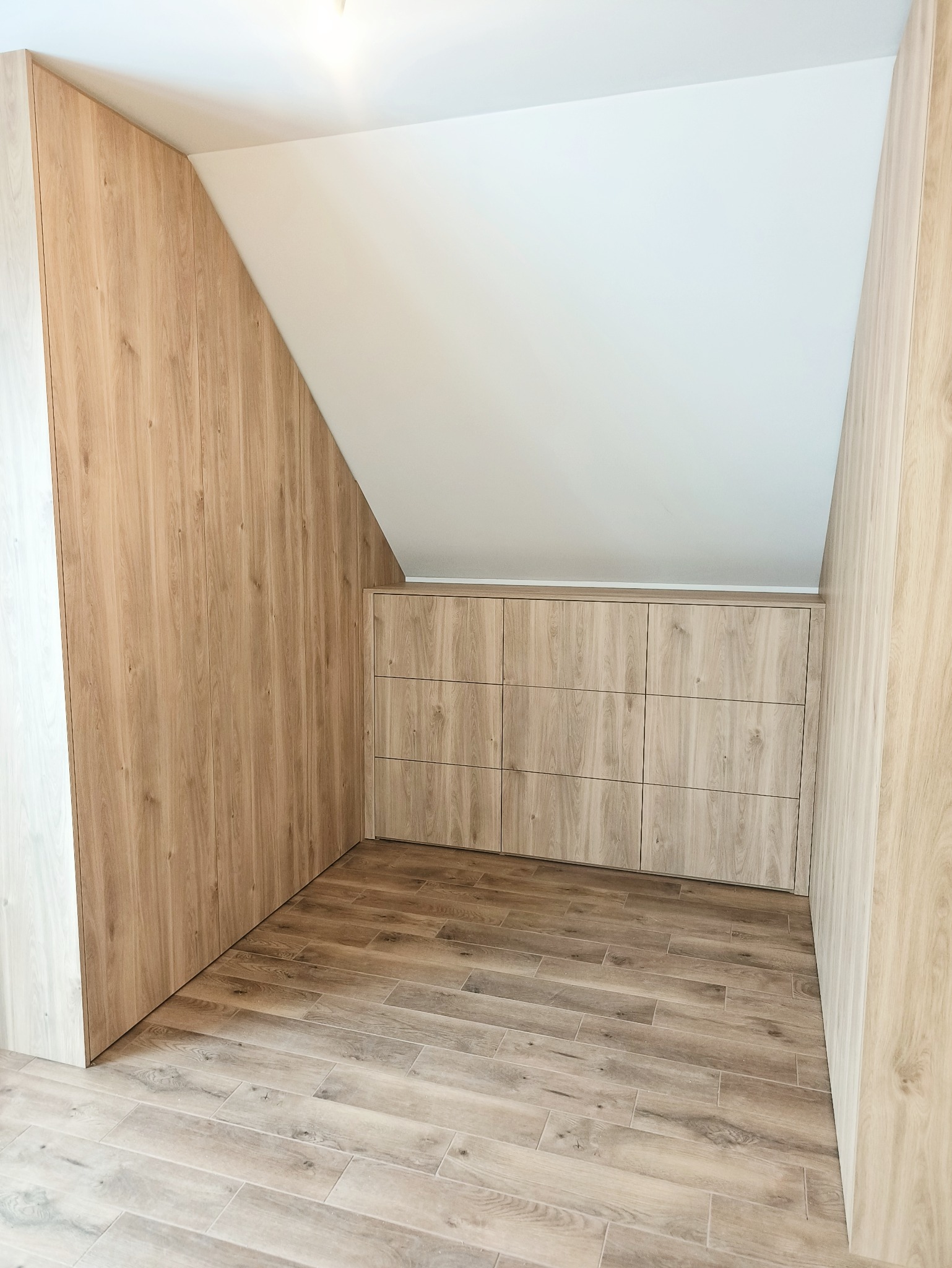 Drewniana zabudowa wnęki na poddaszu z szafkami i panelami ściennymi, dopasowana do skośnego sufitu. Podłoga wyłożona płytkami imitującymi drewno.