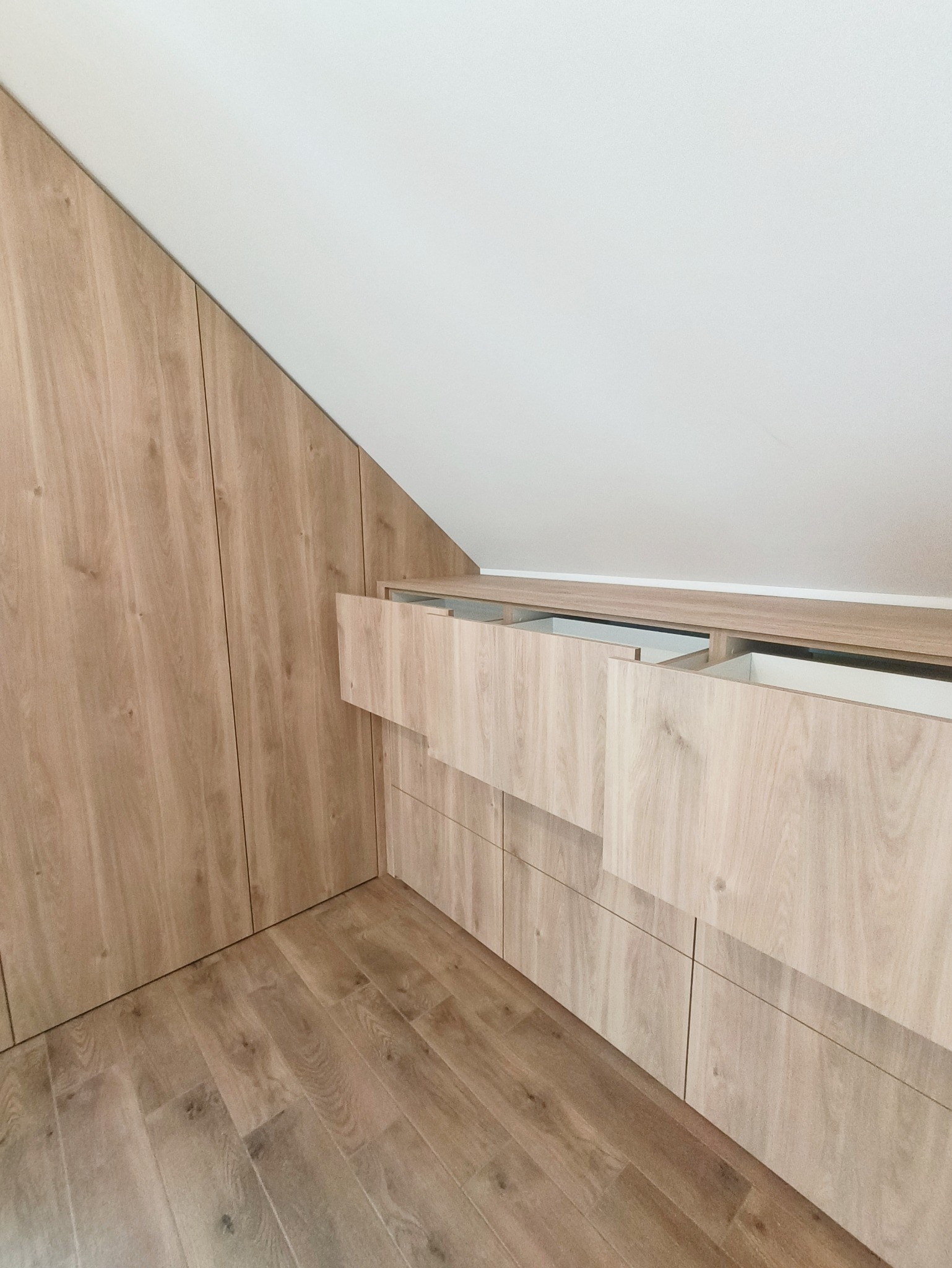 Drewniana szafa wnękowa z szufladami, dopasowana do skosu dachu. Widoczne otwarte szuflady i drewniana podłoga. Ujęcie z dołu, podkreślające wysokość zabudowy.