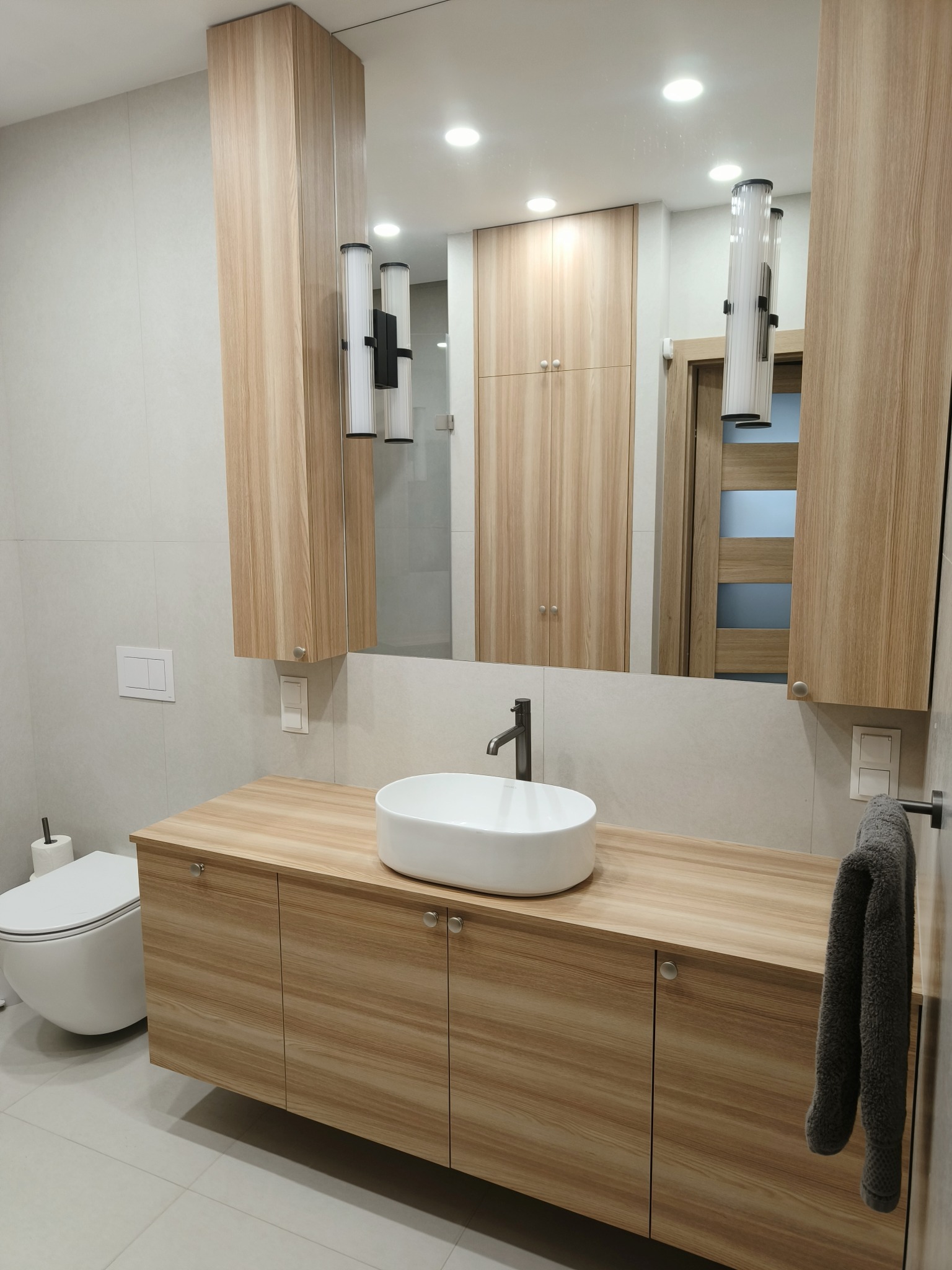 Nowoczesna łazienka z drewnianą szafką pod umywalką, wiszącą toaletą i dużym lustrem z oświetleniem. Widoczne płytki w odcieniach szarości i minimalistyczna armatura.