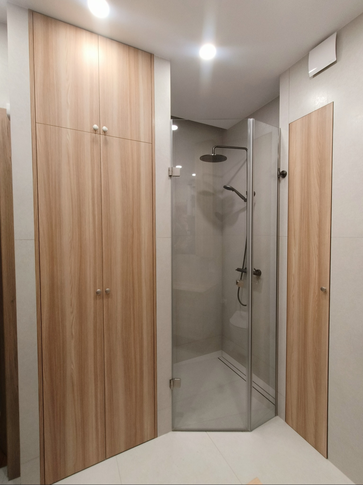 Jasna, drewnopodobna szafa w łazience, obok kabina prysznicowa ze szklanymi drzwiami. Minimalistyczny design, neutralna kolorystyka, oświetlenie sufitowe.