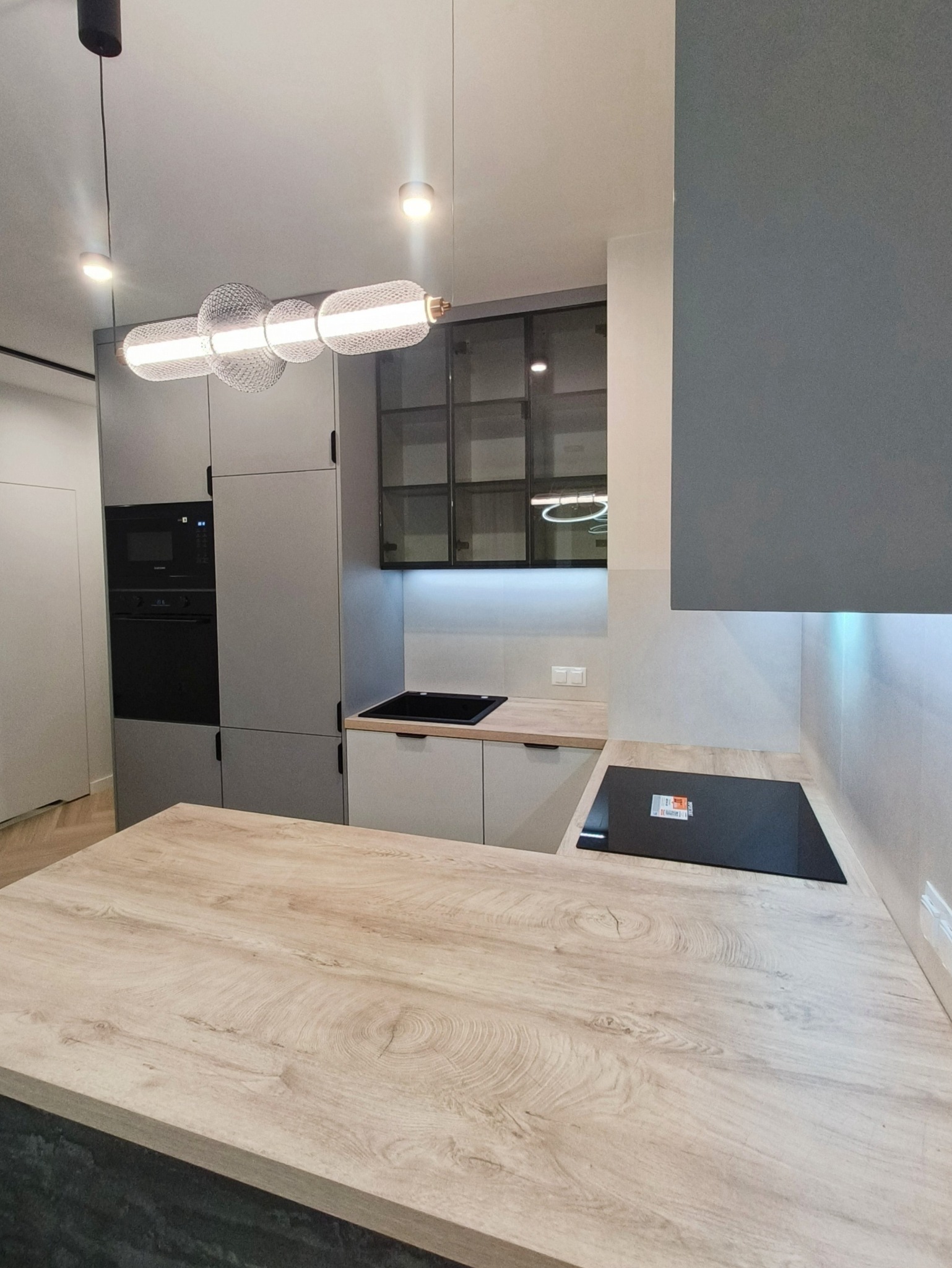 Nowoczesna kuchnia w odcieniach szarości z drewnianym blatem wyspy. Nad blatem wisi designerska lampa. Widoczne zabudowane sprzęty AGD i przeszklone szafki.