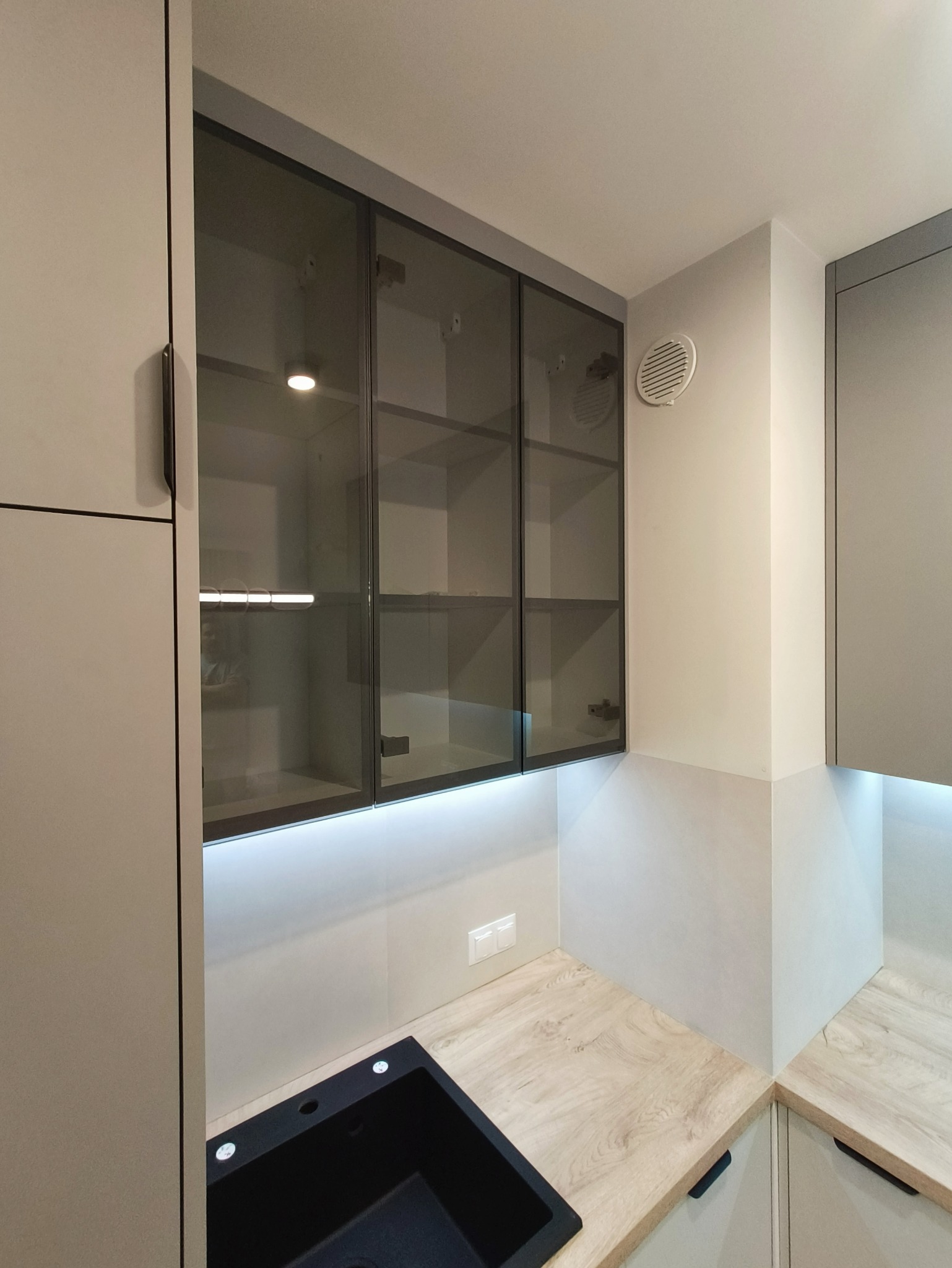 Nowoczesna kuchnia z szarymi szafkami, czarnym zlewem i drewnianym blatem. Górne szafki przeszklone, podświetlane LED. Minimalistyczny design.