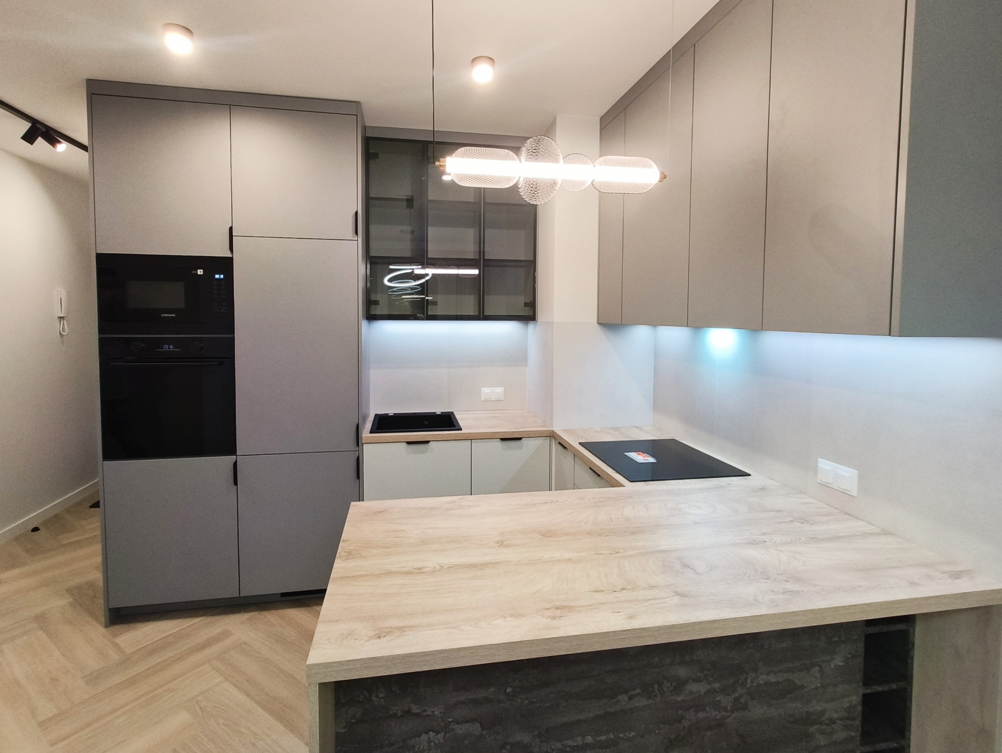 Nowoczesna kuchnia w odcieniach szarości z drewnianym blatem wyspowym i designerską lampą. Minimalistyczne fronty szafek i zabudowany sprzęt AGD.