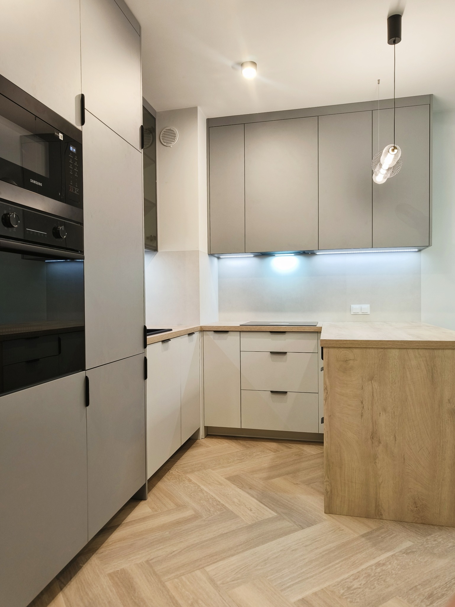 Jasna kuchnia z szarymi frontami, drewnianym blatem i parkietem w jodełkę. Oświetlenie LED pod szafkami. Nowoczesny design z piekarnikiem Samsung i designerską lampą.