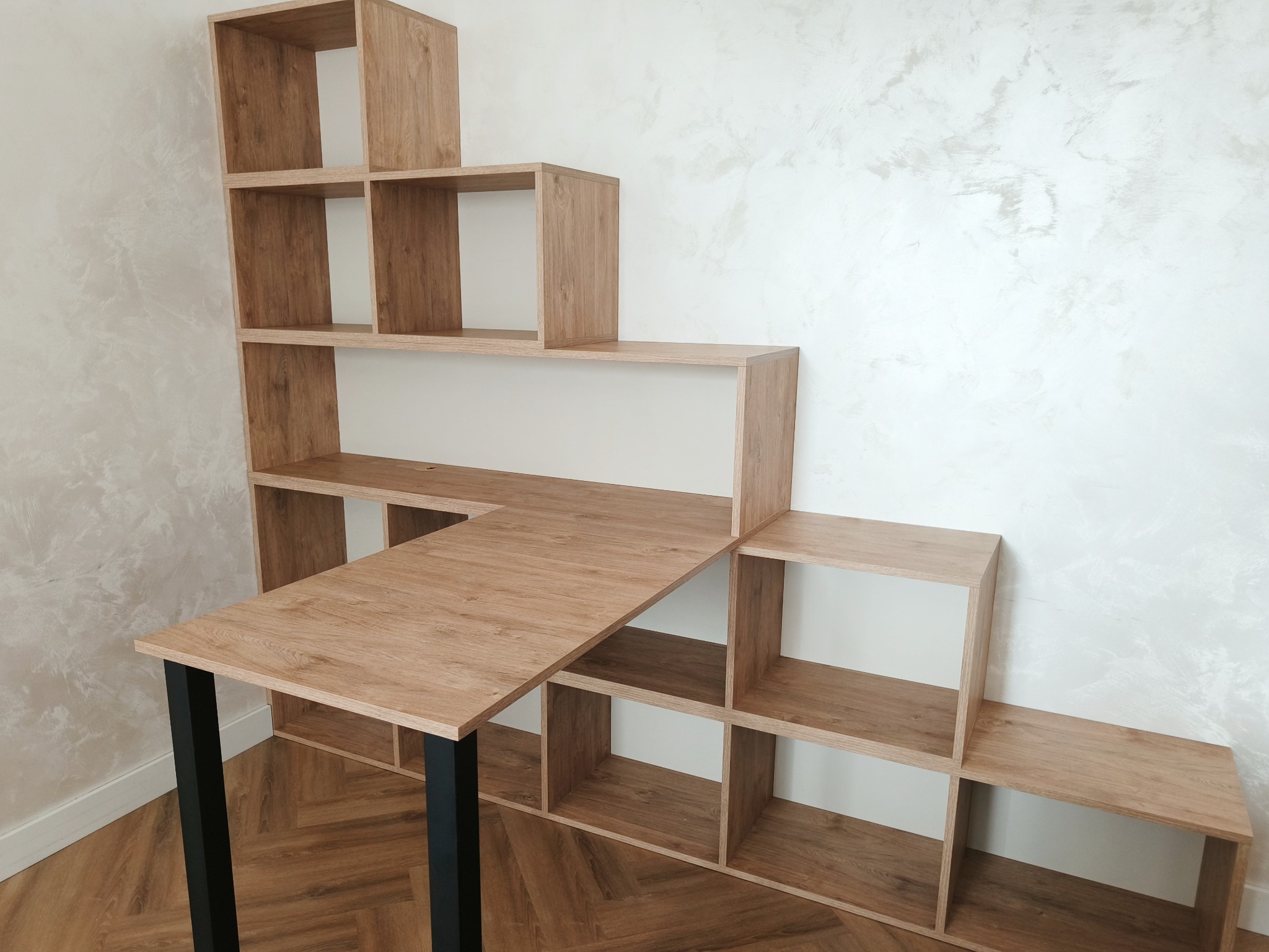 Kreatywna aranżacja: regał schodkowy z blatem biurka na czarnych nogach, drewnopodobny dekor, na tle ściany z efektem betonu i parkietu w jodełkę.