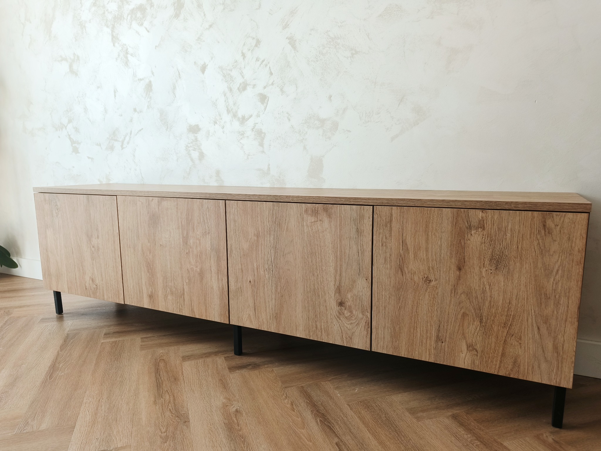 Nowoczesna szafka RTV z drewnianymi frontami i czarnymi nóżkami, na tle ściany z dekoracyjnym tynkiem. Ujęcie z niskiej perspektywy, podkreślające fakturę drewna.