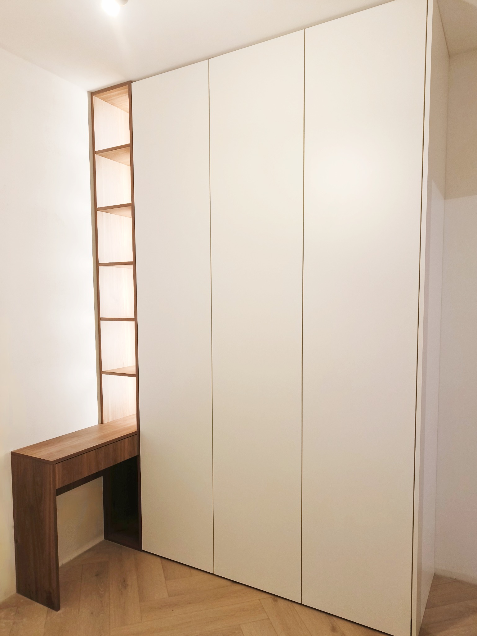 Biała szafa wnękowa z drewnianym biurkiem i regałem. Minimalistyczny design, jasne wnętrze, parkiet w jodełkę. Ujęcie z niskiego kąta.