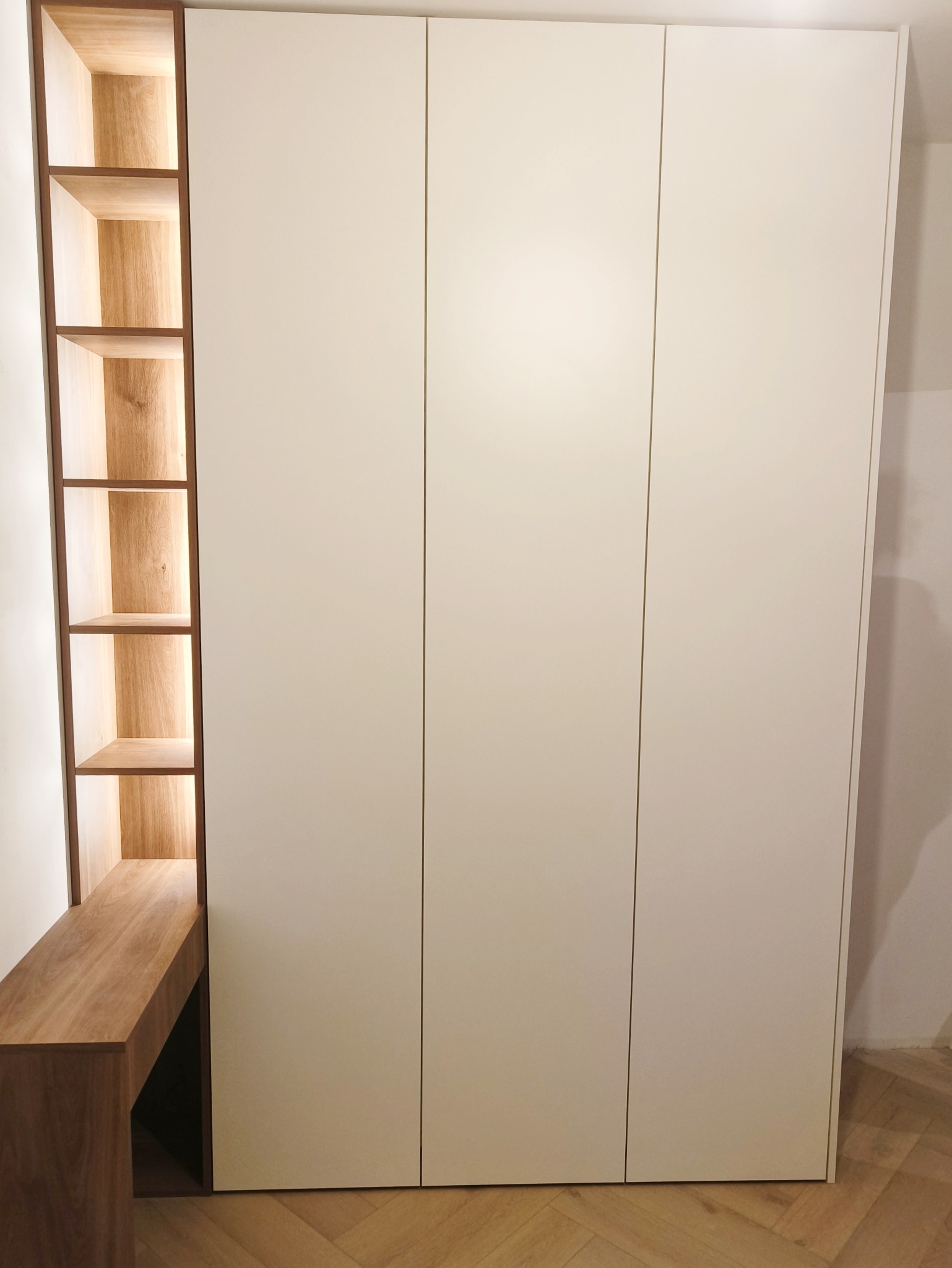 Biała szafa trzydrzwiowa z drewnianym regałem i podświetleniem LED w Ciepłe Pierwsze. Minimalistyczny design, jasne wnętrze, drewniane wykończenie.