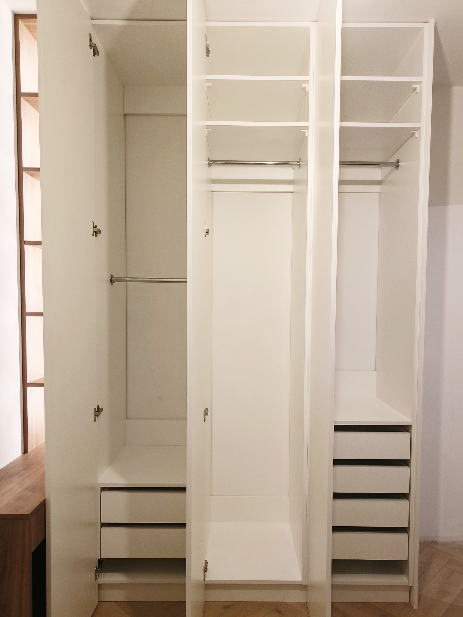 Biała, trzydrzwiowa szafa wnękowa z otwartymi drzwiami, ukazująca półki, drążki i szuflady. Minimalistyczny design, jasne wnętrze, widoczny parkiet na podłodze.