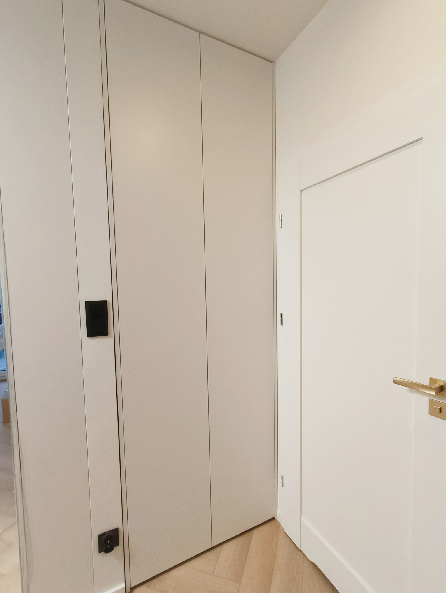 Jasna szafa wnękowa z minimalistycznymi, czarnymi uchwytami, wkomponowana w korytarz z parkietem ułożonym w jodełkę. Obok białe drzwi z złotą klamką.