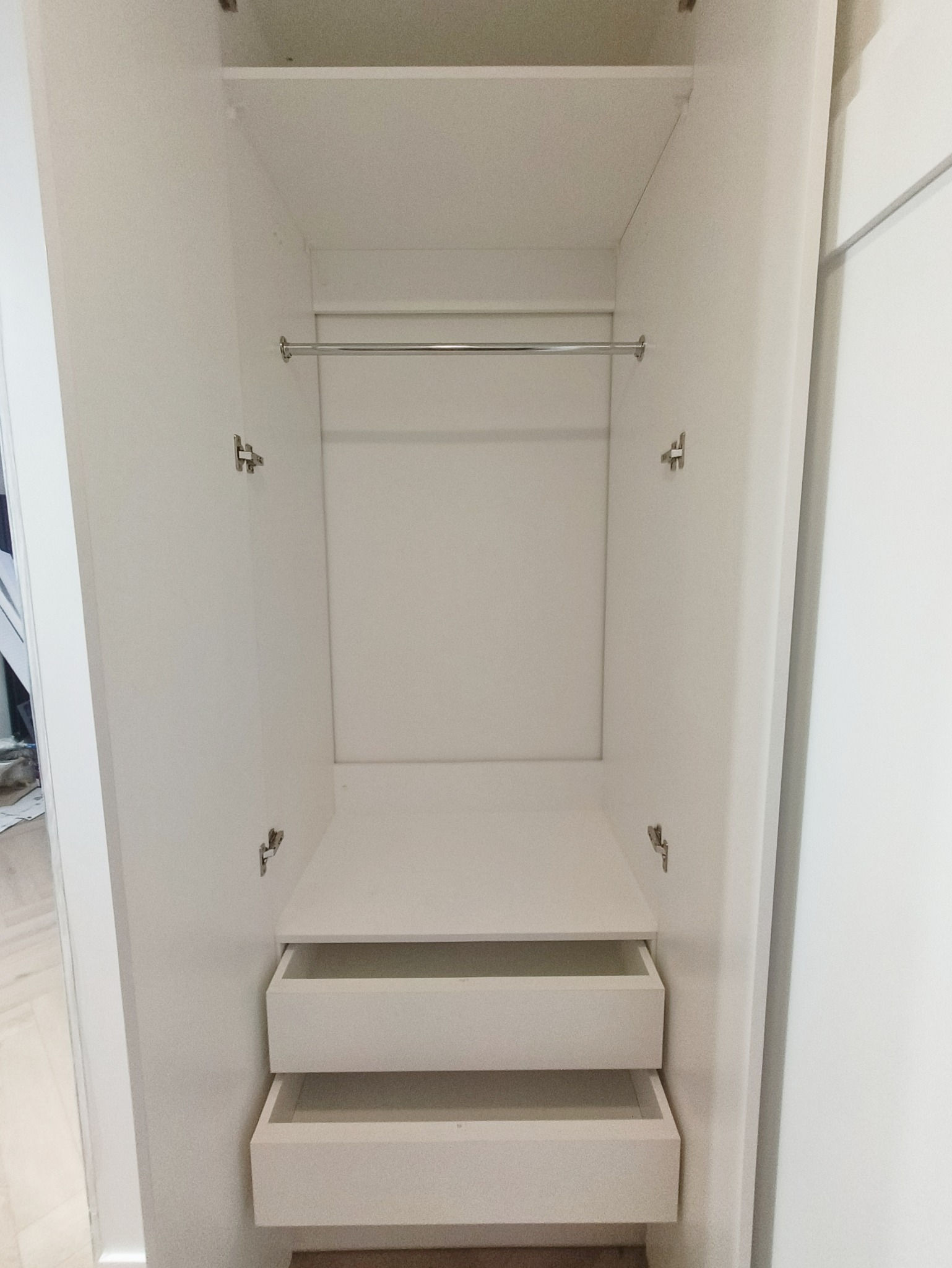 Wnętrze szafy wnękowej z otwartymi drzwiami, widoczne dwie szuflady i drążek na ubrania. Całość w jasnej kolorystyce.