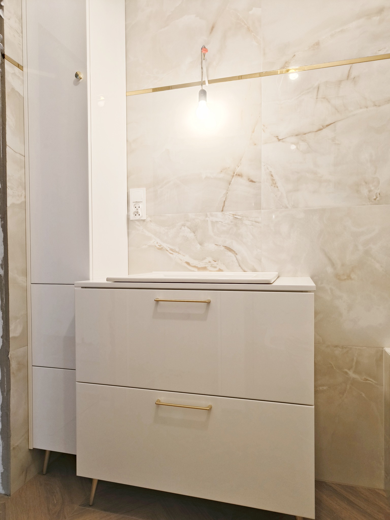 Biała szafka łazienkowa z dwiema szufladami i złotymi uchwytami, na tle marmurowych płytek i wiszącej żarówki. Element zabudowy łazienki.