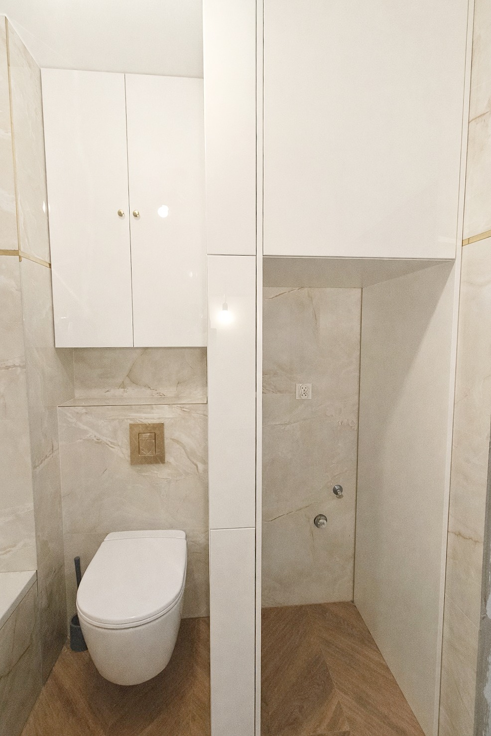 Biała zabudowa łazienkowa z szafkami nad toaletą i wnęką prysznicową. Beżowe płytki imitujące marmur, drewnopodobna podłoga ułożona w jodełkę.