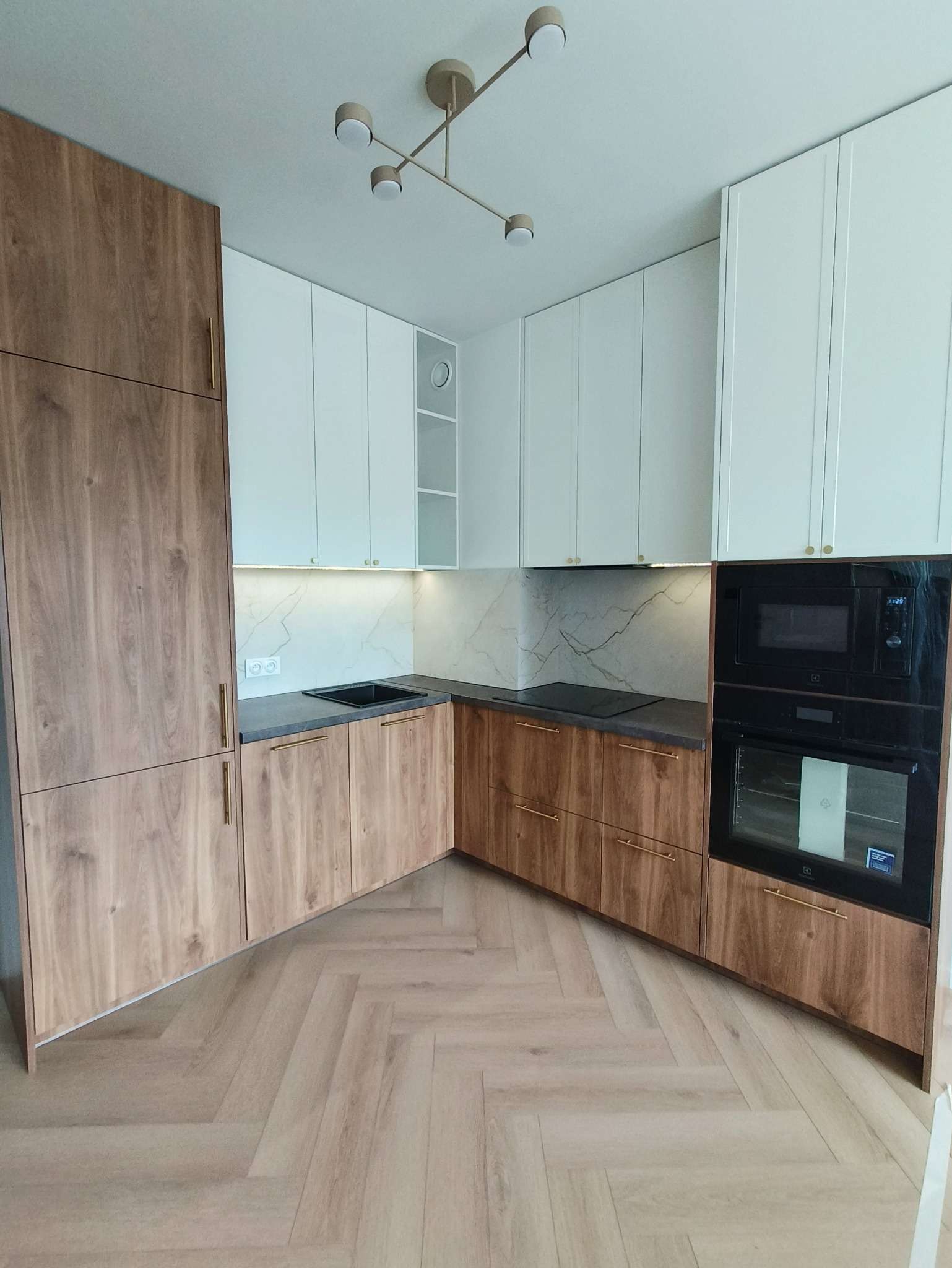 Nowoczesna kuchnia z drewnianymi frontami i białymi szafkami wiszącymi, czarnym zlewem i sprzętem AGD w zabudowie. Parkiet w jodełkę na podłodze.