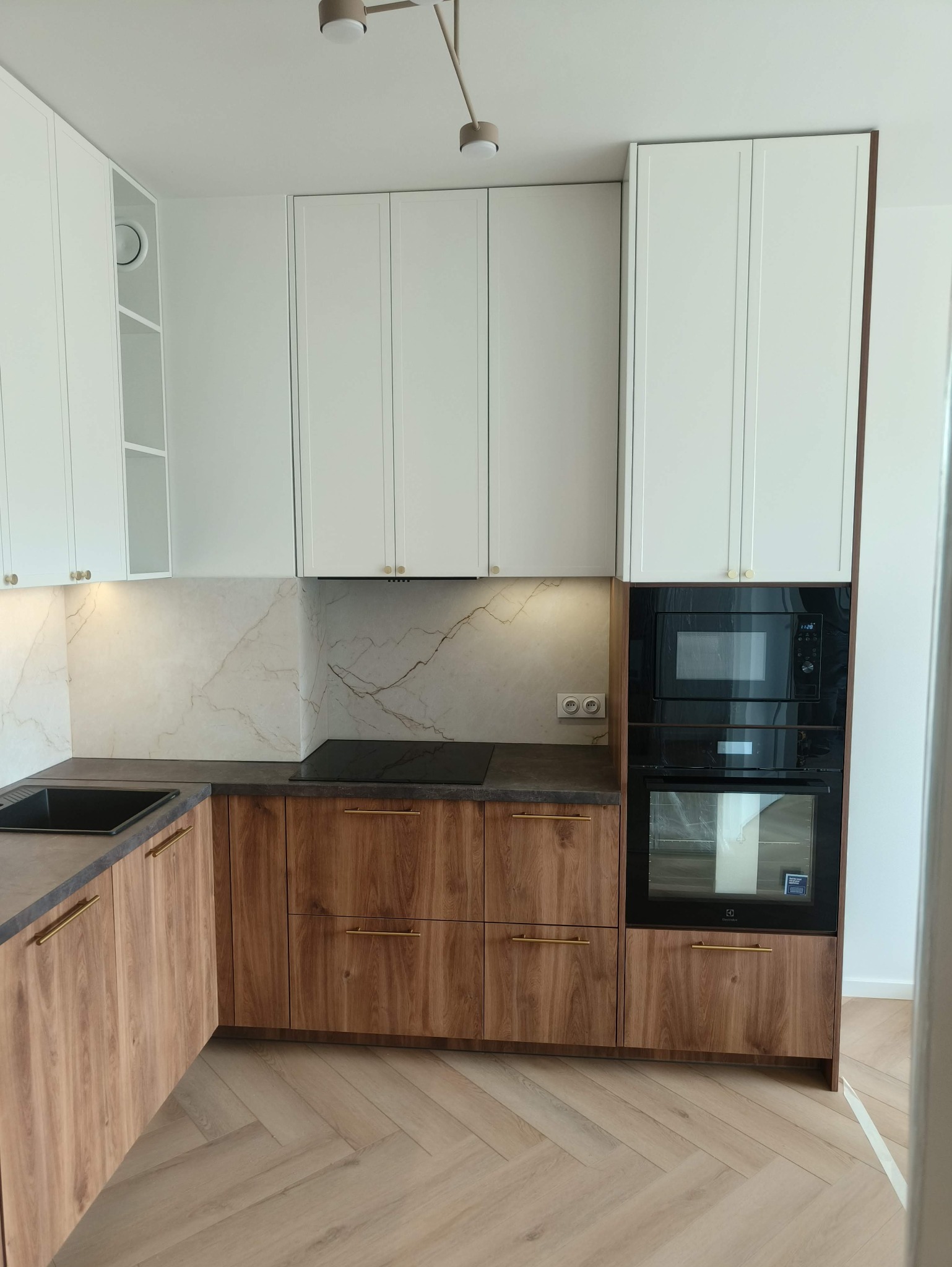 Nowoczesna kuchnia z białymi szafkami górnymi i drewnopodobnymi dolnymi, złote uchwyty, marmurowy backsplash, czarny piekarnik i płyta, parkiet w jodełkę.