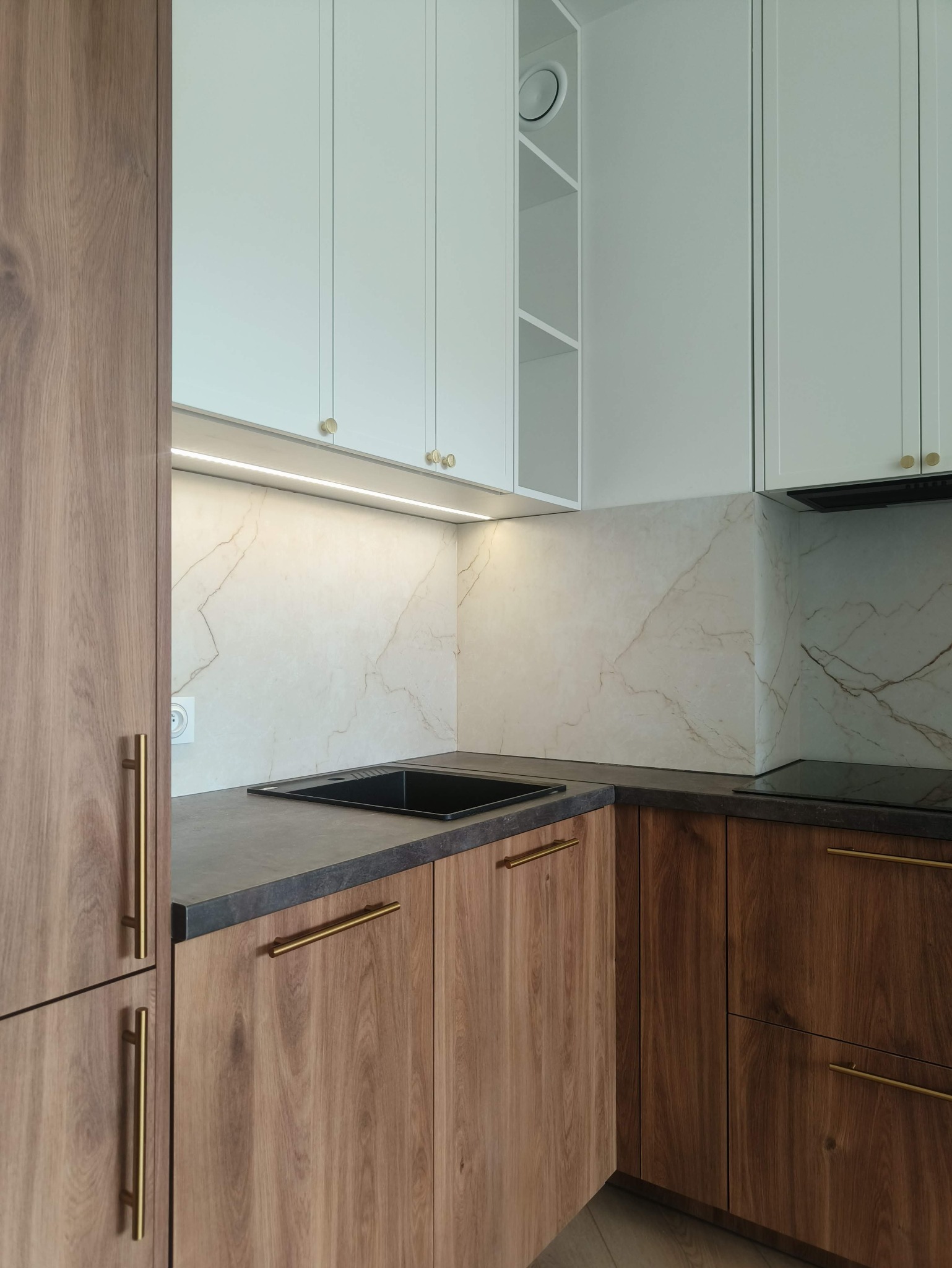 Nowoczesna zabudowa kuchenna w Ciepłe Pierwsze. Kontrastujące, drewniane fronty szafek dolnych i białe górne. Uwagę zwracają złote uchwyty i marmurowy wzór na ścianie.