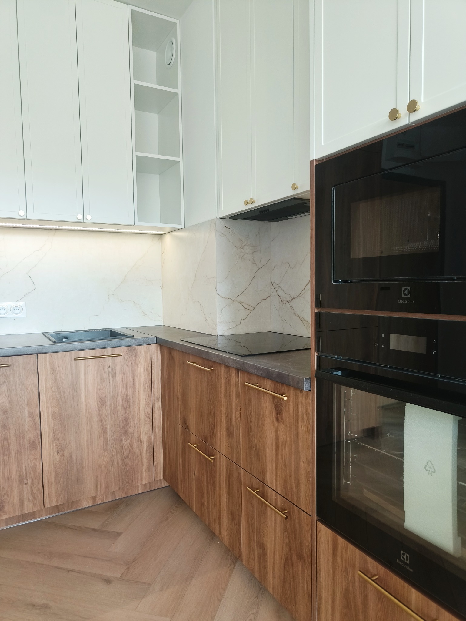 Nowoczesna kuchnia z drewnianymi frontami szafek dolnych i białymi górnymi, złote uchwyty, marmurowy backsplash i zabudowany sprzęt AGD Electrolux.