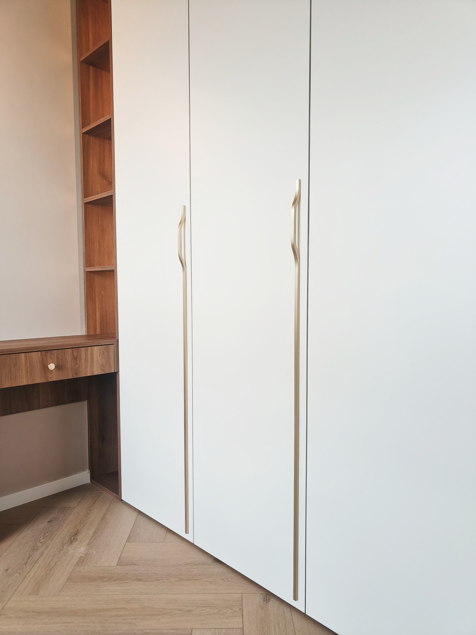 Biała szafa na wymiar z złotymi uchwytami i drewnianym biurkiem z półkami w Ciepłe Pierwsze. Parkiet w jodełkę. Minimalistyczny design, jasne wnętrze.