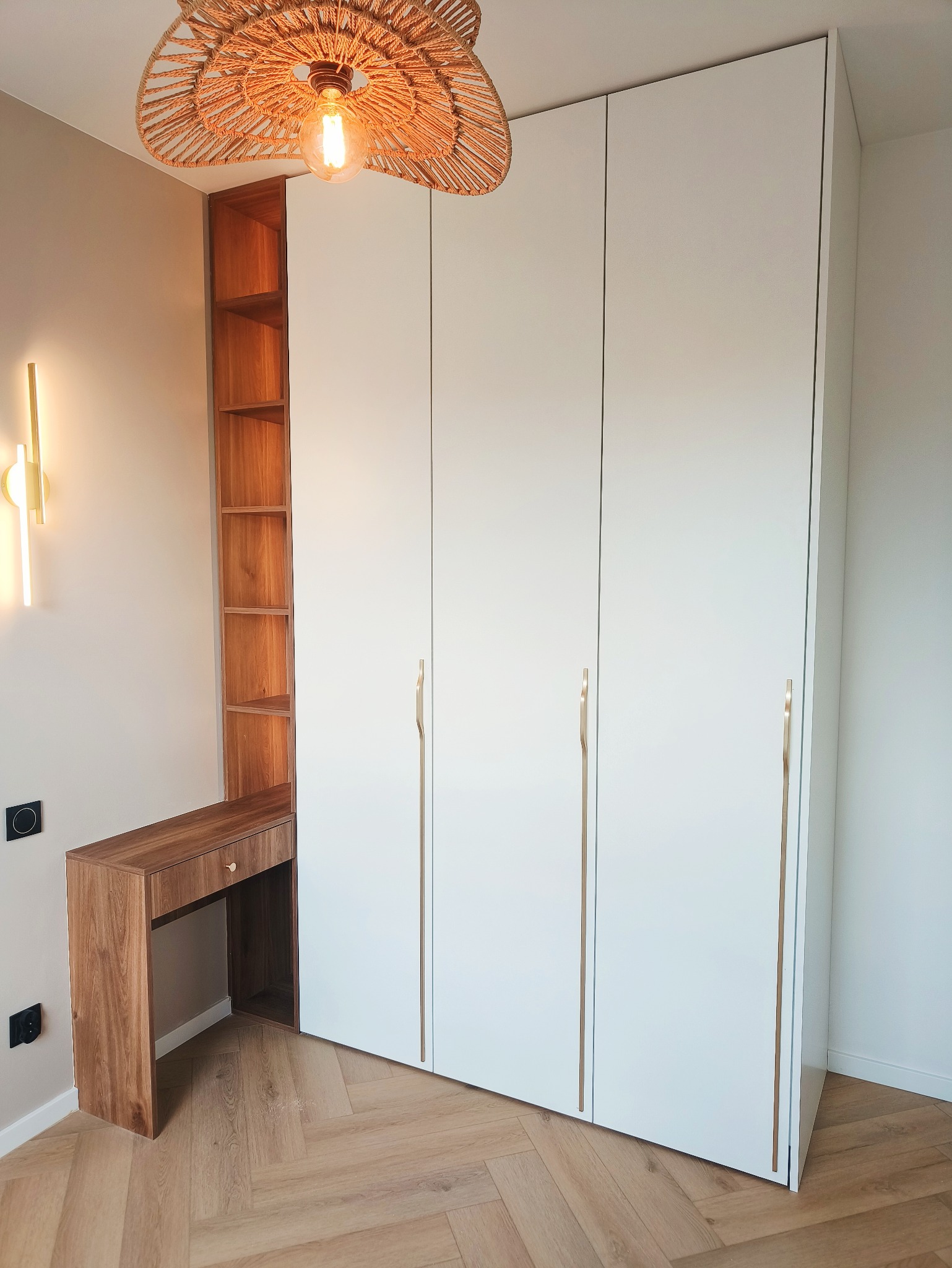 Biała szafa na wymiar z drewnianym biurkiem i regałem, złote uchwyty, parkiet w jodełkę, lampa z abażurem z rafii. Nowoczesny design, ciepłe oświetlenie.