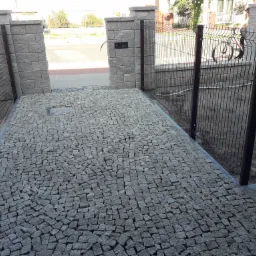 Ukończone wejście na posesję z czarnego ogrodzenia panelowego, kamiennej kostki brukowej i słupków z szarej cegły, z widocznym przechodzącym rowerzystą w tle.