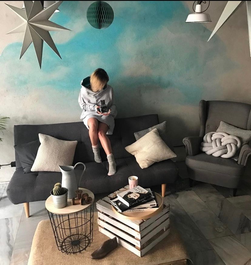 Ściana z efektem akwareli w odcieniach błękitu, na tle której siedzi osoba z aparatem, otoczona elementami wystroju wnętrz w stylu skandynawskim.