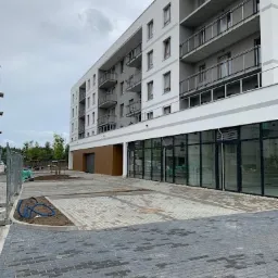 Nowo ułożona, szara kostka brukowa przed nowoczesnym, białym budynkiem apartamentowym z balkonami, widoczne przygotowane miejsce pod parking, częściowo ogrodzone tymczasowym płotem.