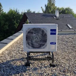 Biała jednostka zewnętrzna klimatyzacji Haier na dachu pokrytym żwirem, na metalowym stelażu. W tle budynek z ciemną dachówką i zieleń drzew.