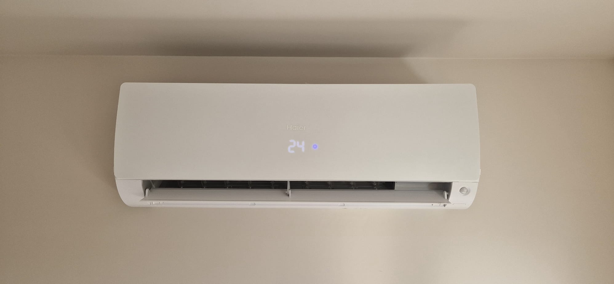 Biała klimatyzacja Haier na ścianie, wyświetlacz pokazuje 24 stopnie. Widok z przodu, minimalistyczny design, subtelne oświetlenie, domowe wnętrze.