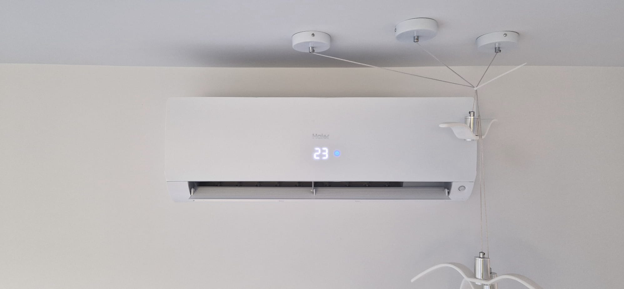 Biały klimatyzator Haier na ścianie, wyświetla temperaturę 23 stopnie. Nietypowe oświetlenie sufitowe z trzema punktami świetlnymi.