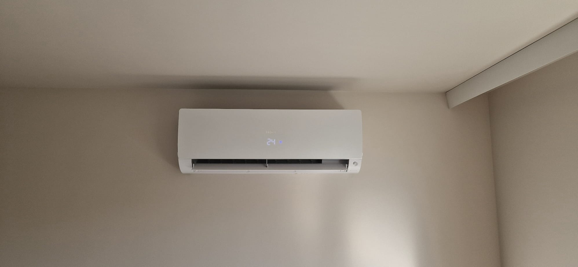 Biały klimatyzator Haier na beżowej ścianie, wyświetlający temperaturę 24 stopnie. Widoczne detale konstrukcyjne i subtelne oświetlenie.