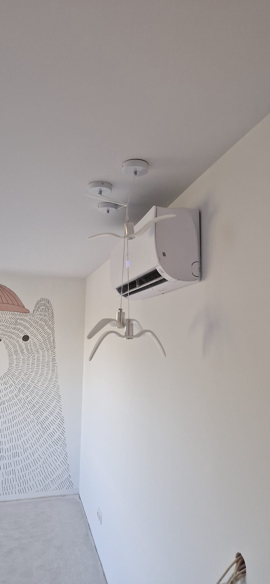 Biała klimatyzacja ścienna w pokoju dziecięcym z lampą w kształcie ptaków. Widoczny fragment ściany z rysunkiem misia. Minimalistyczny, nowoczesny design.